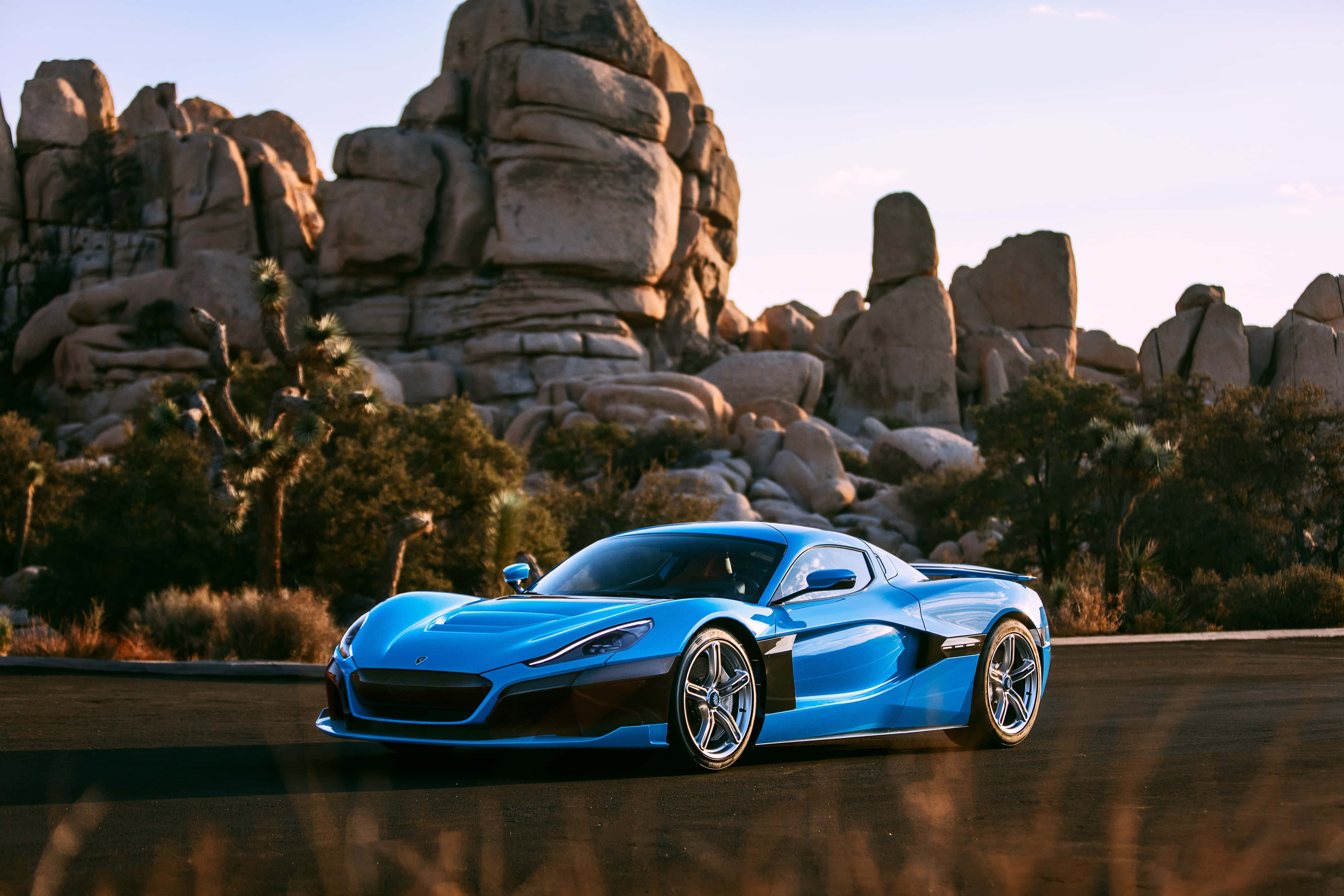 Rimac 桌布 壁紙 — 一輛充滿活力的藍色跑車在金色日落光芒下的沙漠公路上閃閃發光，周圍是高聳的岩層和稀疏的沙漠植被。