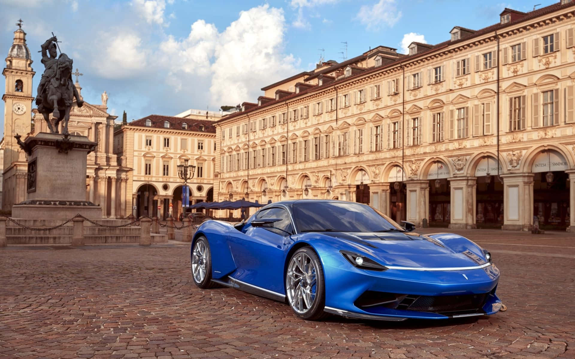 Pininfarina 桌布 壁紙 — 一輛充滿活力的藍色跑車在鵝卵石廣場上閃閃發光，旁邊是歷史悠久的騎馬雕像和華麗的建築，天空部分多雲。