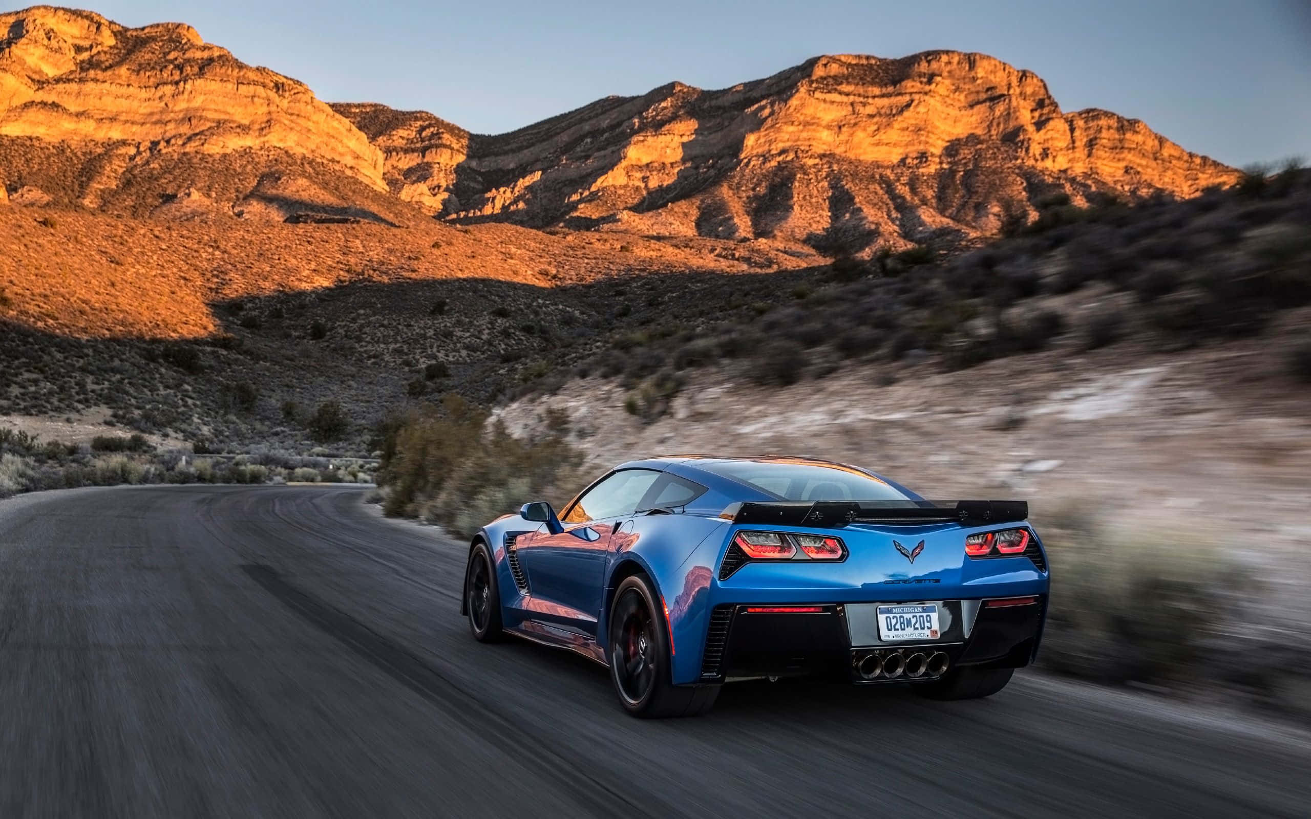 Chevrolet Corvette Z06 桌布 壁紙 — 一輛充滿活力的藍色跑車沿著蜿蜒的沙漠公路疾馳，崎嶇的山脈沐浴在金色的日落光芒中。