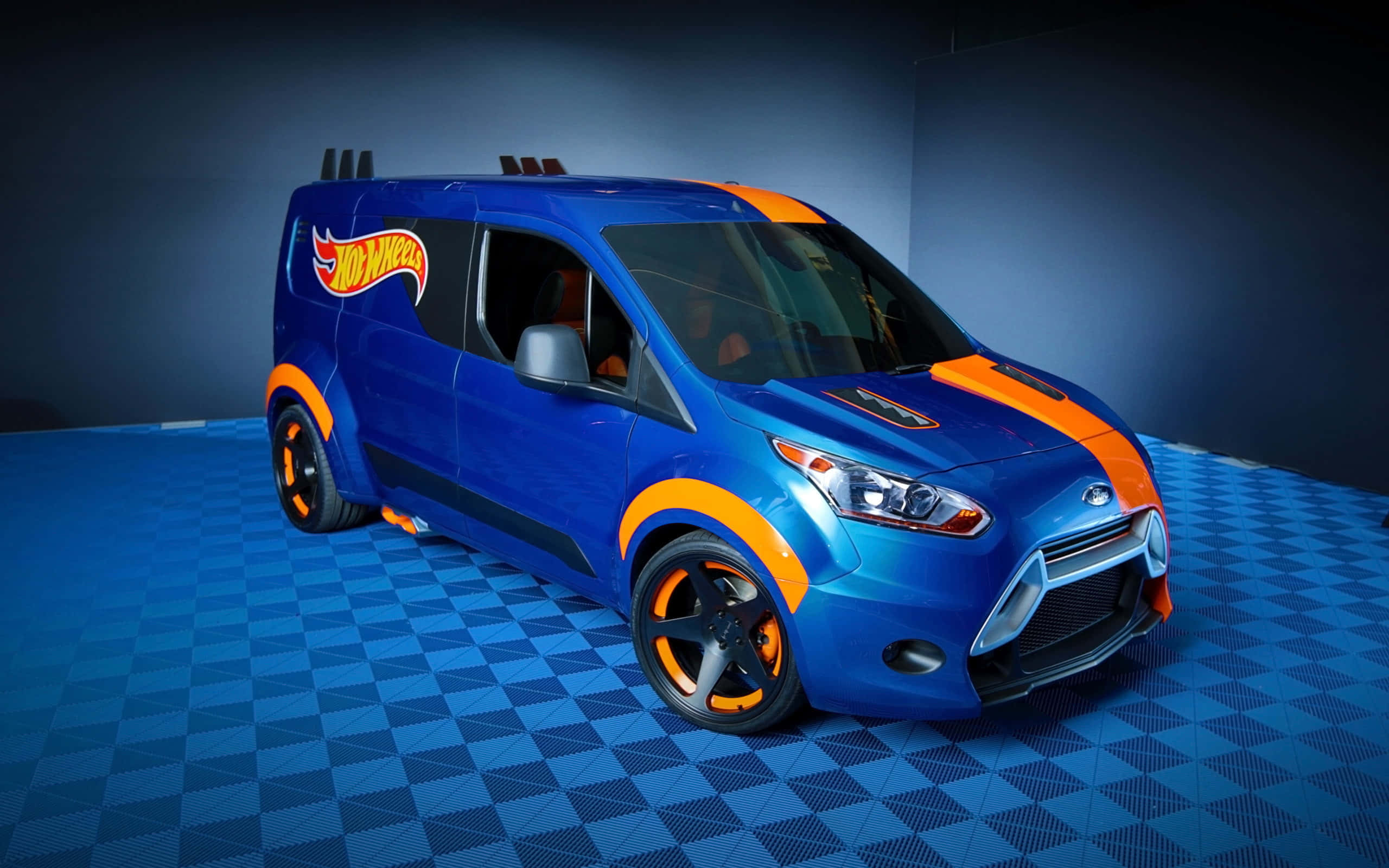 Ford Transit 桌布 壁紙 — 一輛充滿活力的藍色風火輪貨車，帶有橙色點綴和定制輪圈，在工作室燈光下停放在格子地板上。