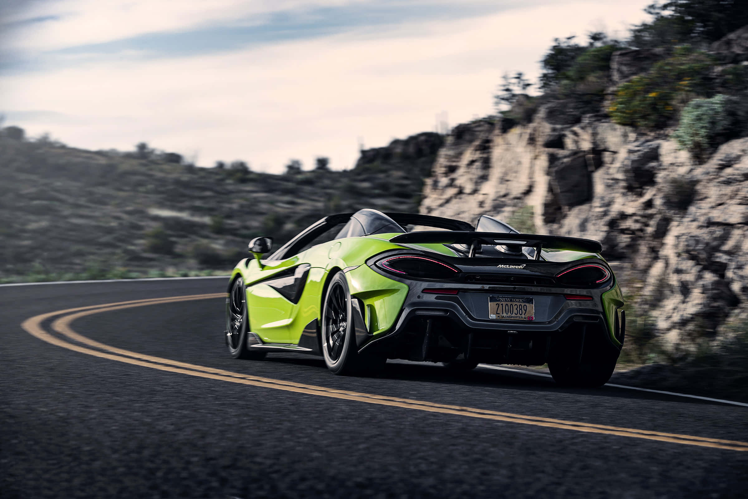 Mclaren 600lt Spider 桌布 壁紙 — 一輛充滿活力的青檸綠色McLaren跑車在蜿蜒的山路上飛馳，兩旁是岩石峭壁和稀疏的植被，天空多雲。