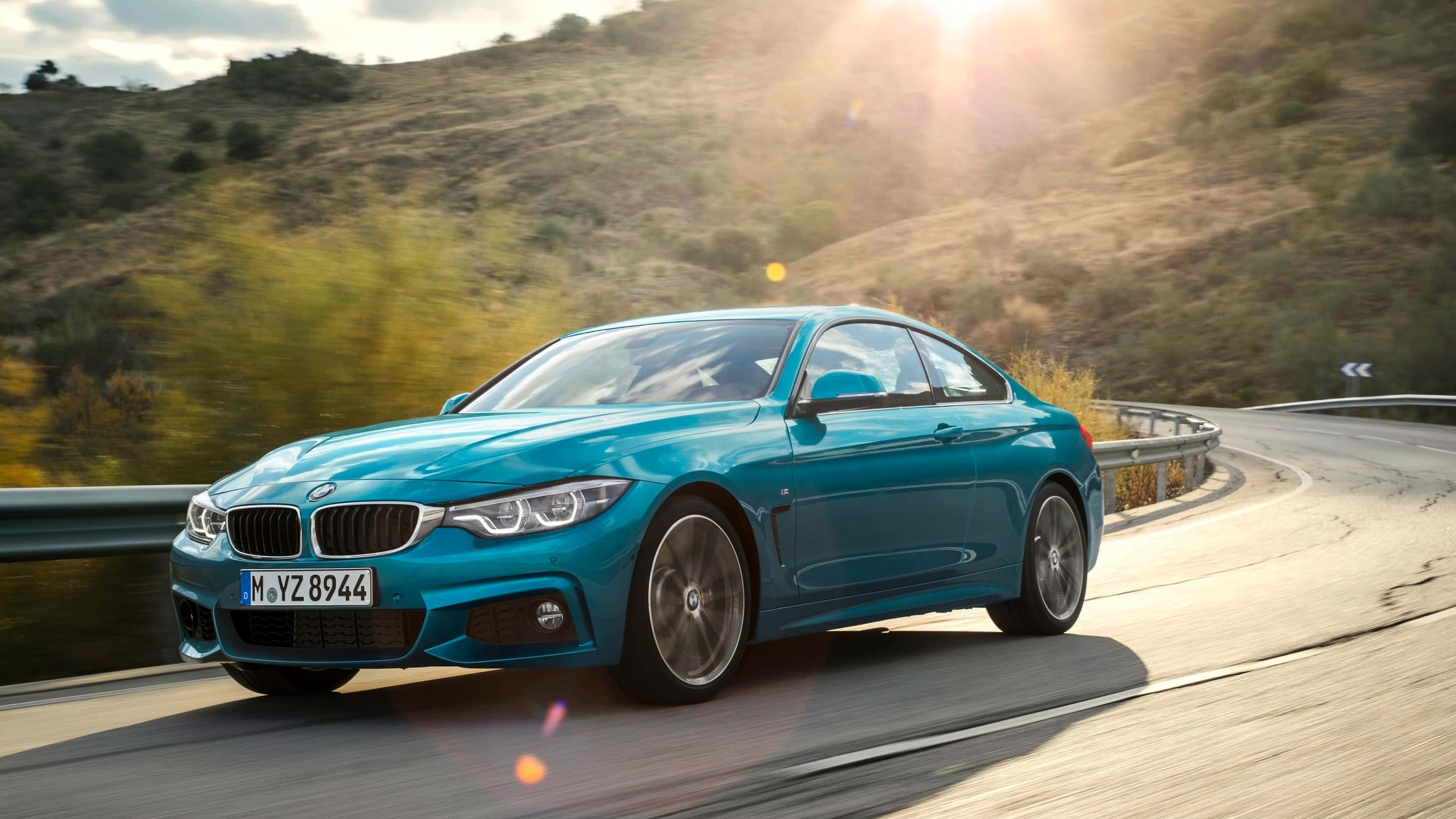 Bmw 4 Series 桌布 壁紙 — 一輛充滿活力的青綠色BMW雙門跑車在蜿蜒的山路上疾馳，沐浴在金色的夕陽光芒中。