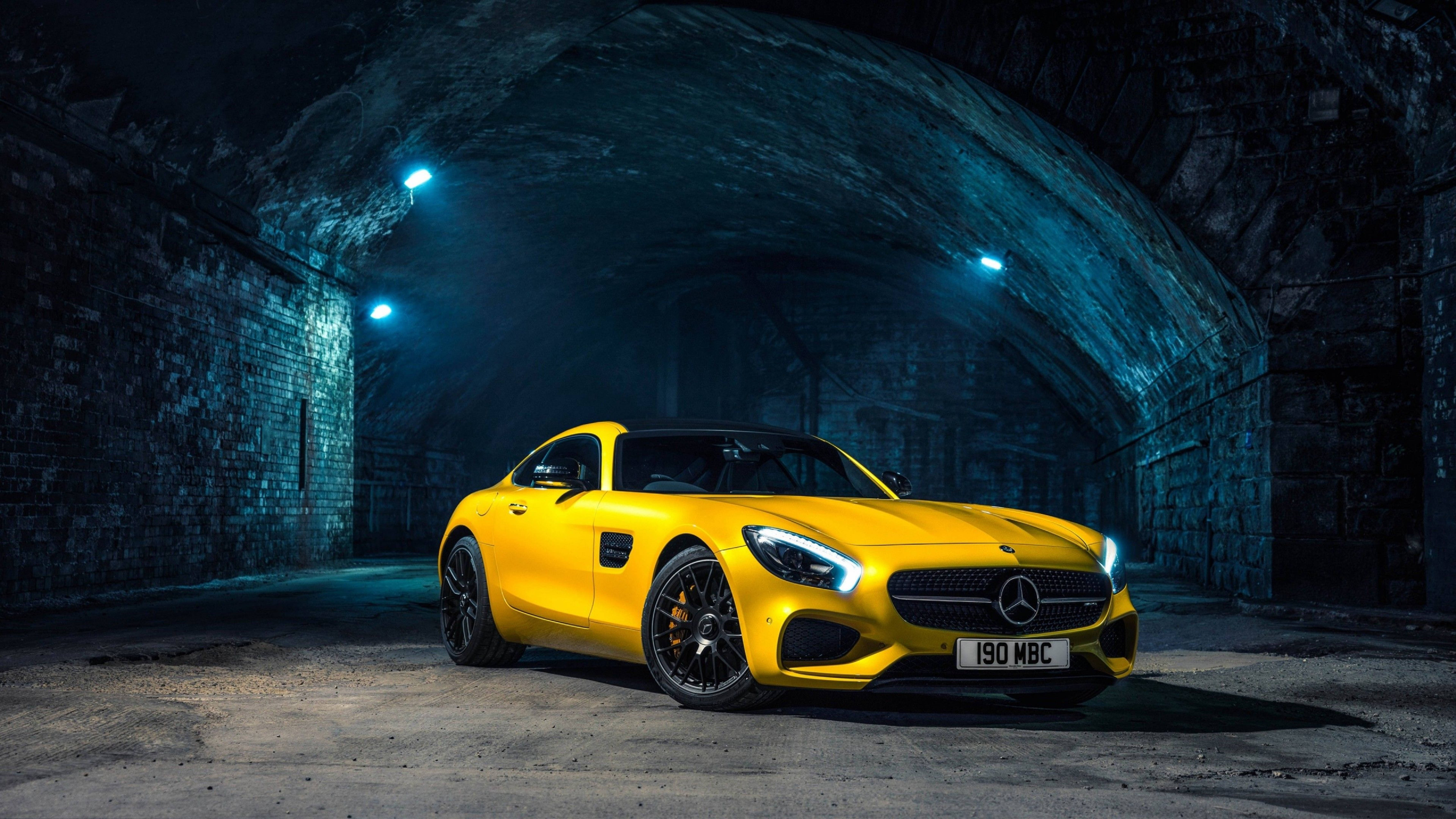 cars 桌布 壁紙 — 一輛充滿活力的黃色Mercedes-AMG GT跑車在藍色隧道燈光下，襯托著粗獷的石拱門閃閃發光。