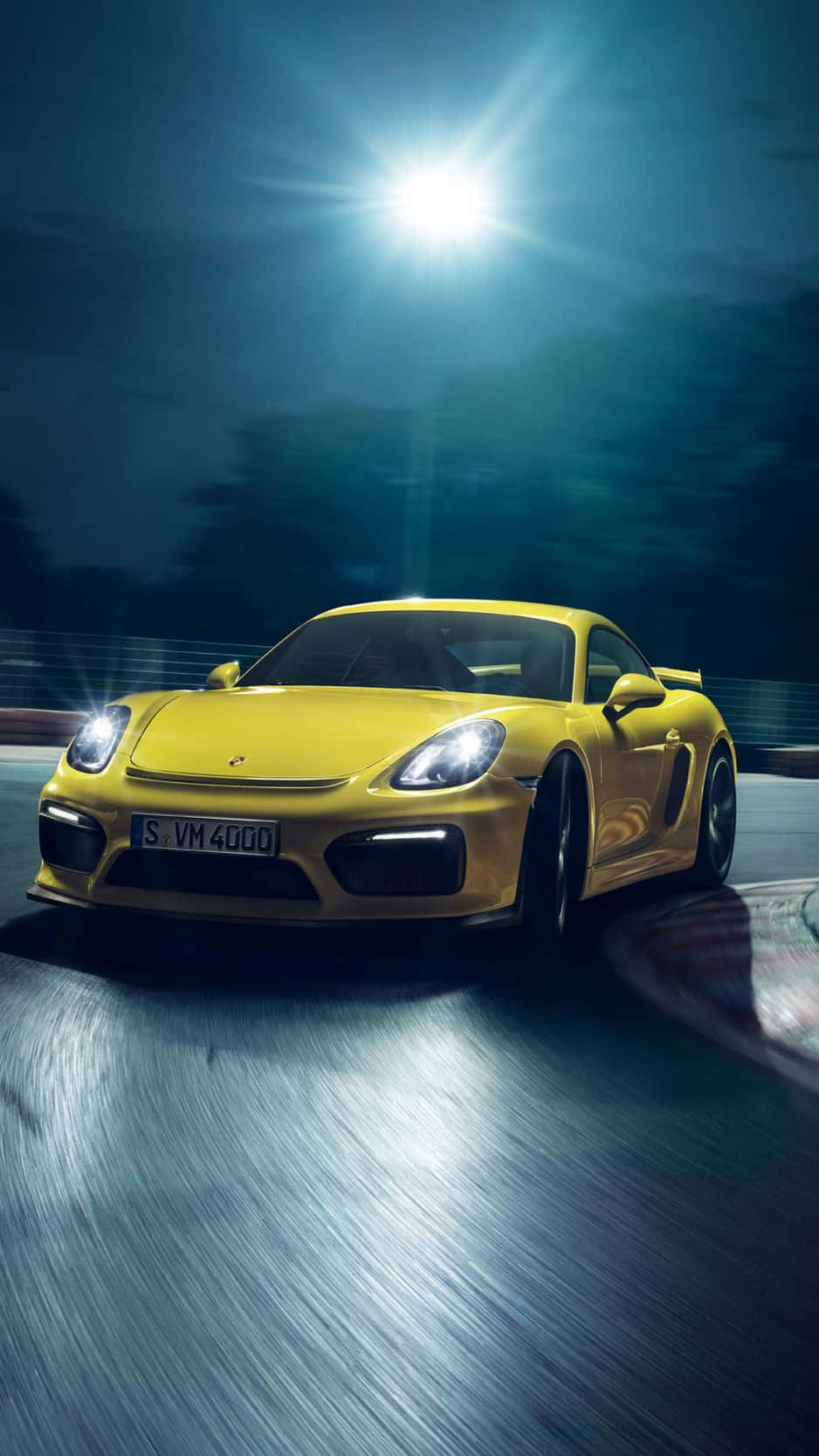 Porsche iPhone 桌布 壁紙 — 一輛充滿活力的黃色保時捷 Cayman 在夜間模糊的賽道上，於明亮的聚光燈下奔馳。