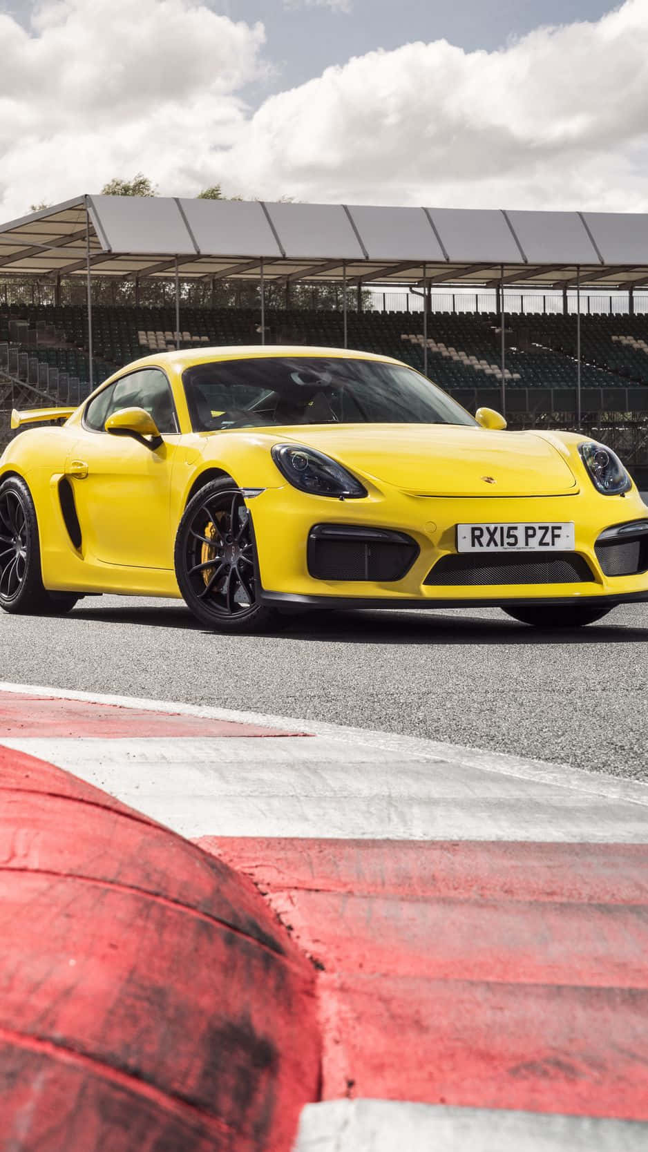 Porsche iPhone 桌布 壁紙 — 一輛充滿活力的黃色保時捷 Cayman GT4 在陰天之下，於設有紅白路緣的賽道上奔馳。