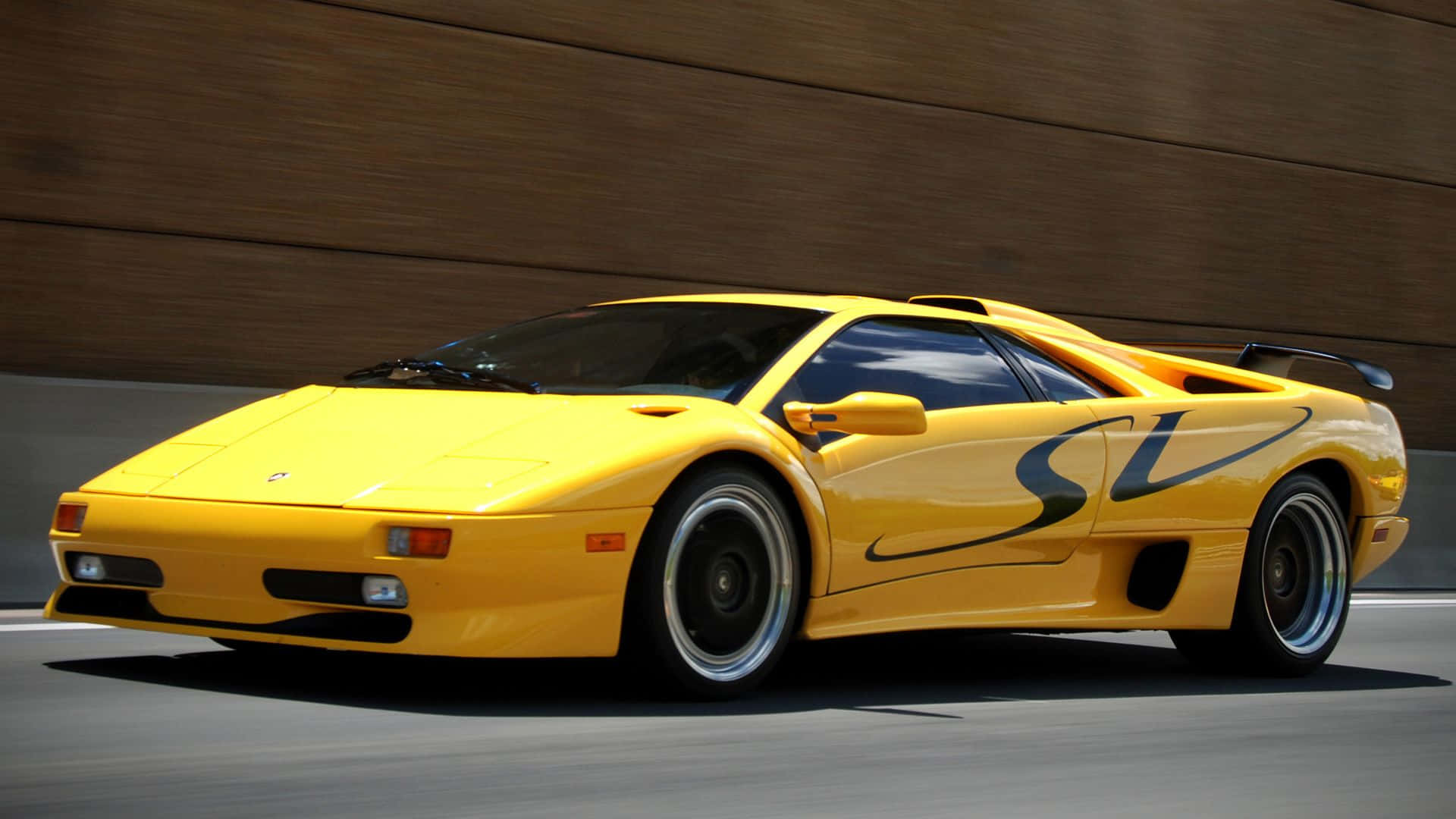 Lamborghini Diablo 桌布 壁紙 — 一輛充滿活力的黃色林寶堅尼 Diablo SV 在道路上奔馳，帶有動態模糊效果，背景為木牆。