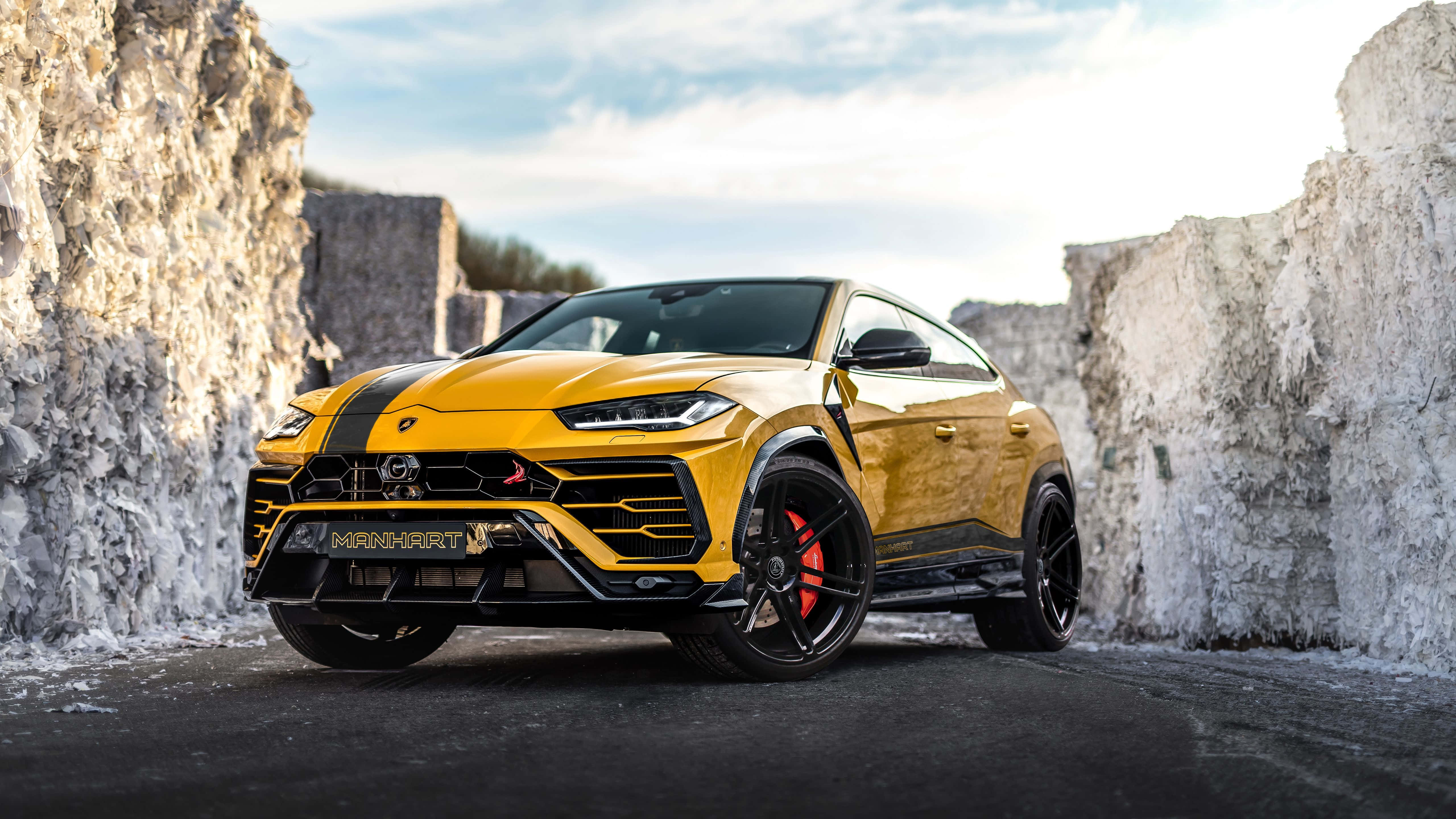 Lamborghini Urus 桌布 壁紙 — 一輛充滿活力的黃色林寶堅尼 Urus，帶有黑色飾件和紅色制動卡鉗，停泊在聳立的白色石崖之間，天空部分多雲。