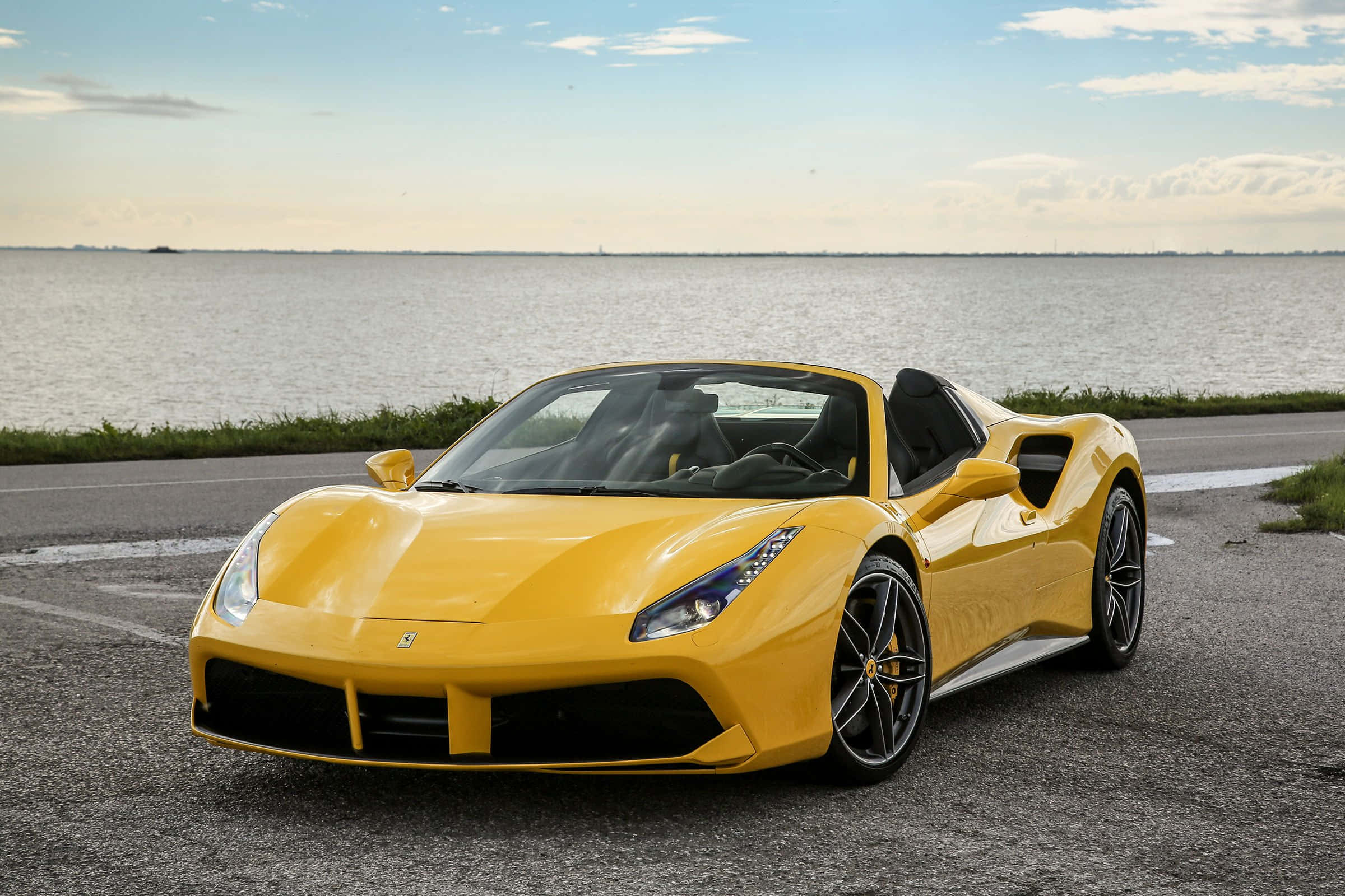 Ferrari 488 Spider 桌布 壁紙 — 一輛充滿活力的黃色法拉利488 Spider停泊在鋪砌的道路旁，旁邊是平靜的水面，天空部分多雲。