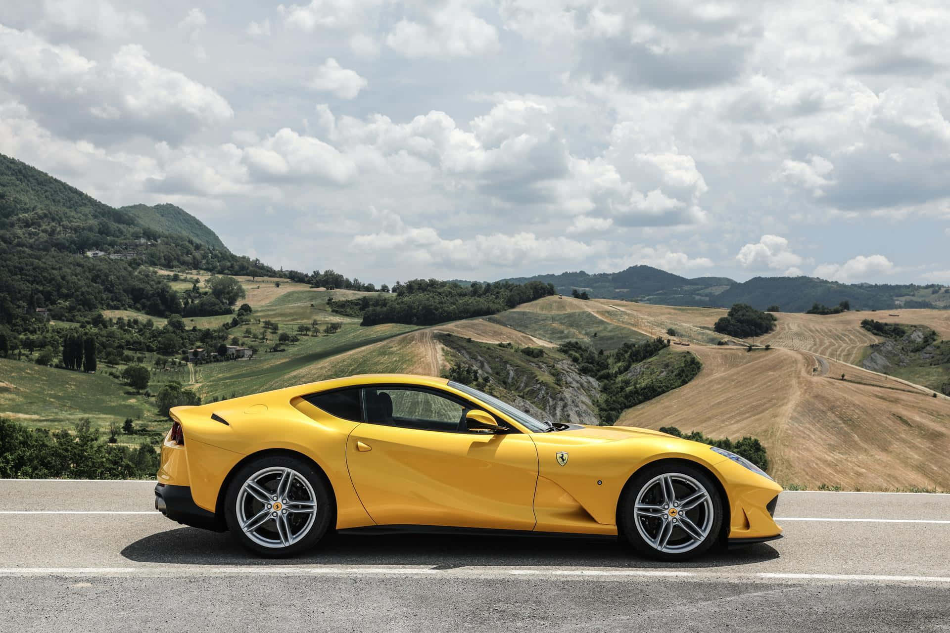 Ferrari 812 Superfast 桌布 壁紙 — 一輛充滿活力的黃色法拉利跑車沿著蜿蜒的道路，在陰雲密布的天空下穿過起伏的綠色和金色山丘。