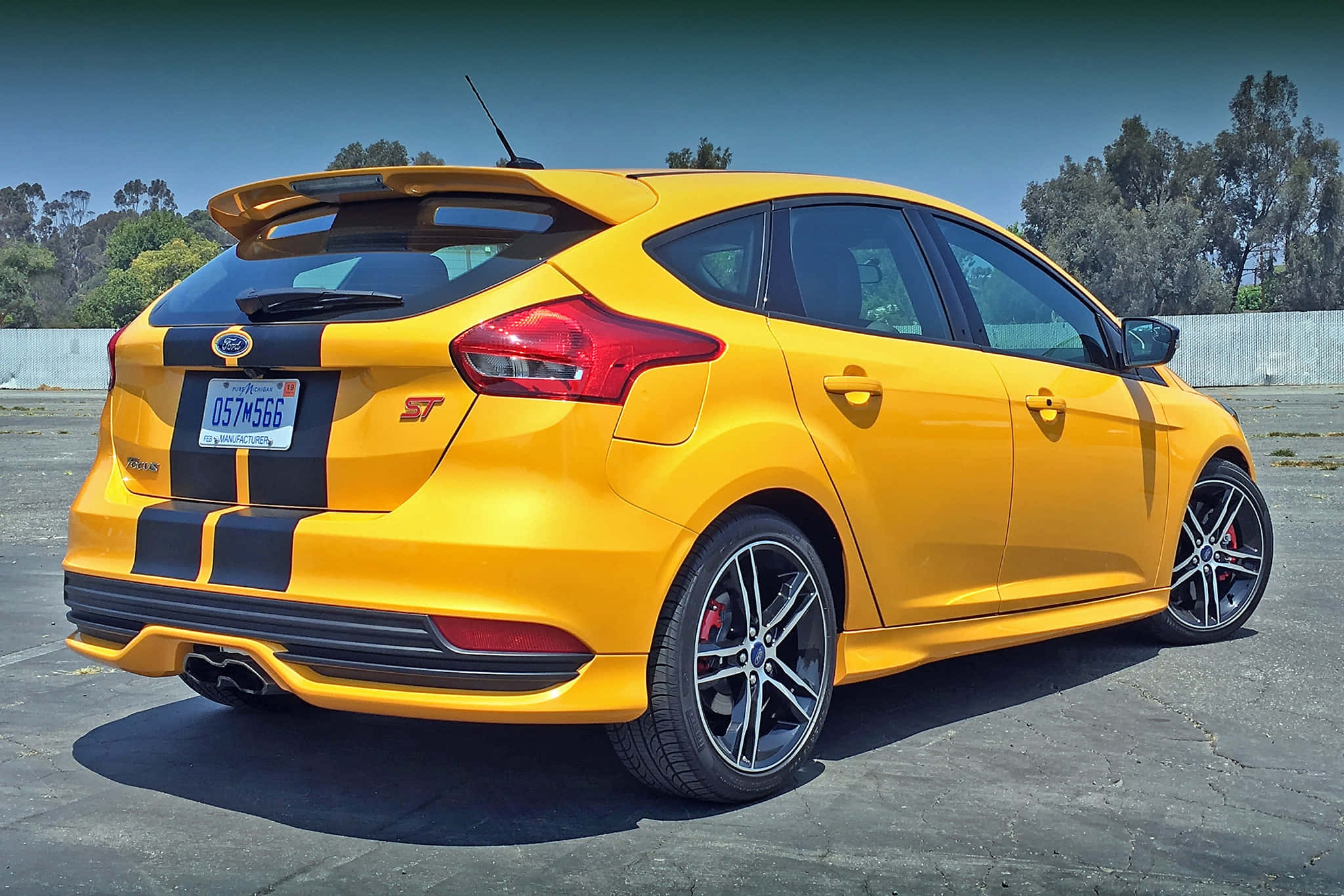Ford Focus 桌布 壁紙 — 一輛充滿活力的黃色福特福克斯ST，帶有黑色賽車條紋，停在晴朗藍天下的瀝青路面上。