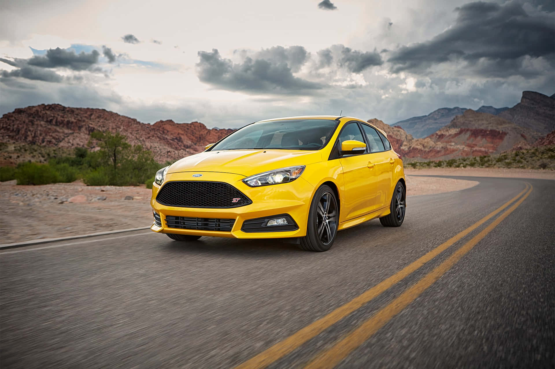 Ford Focus 桌布 壁紙 — 一輛充滿活力的黃色福特福克斯ST在戲劇性的多雲天空下沿著沙漠道路飛馳，背景是紅色岩層。