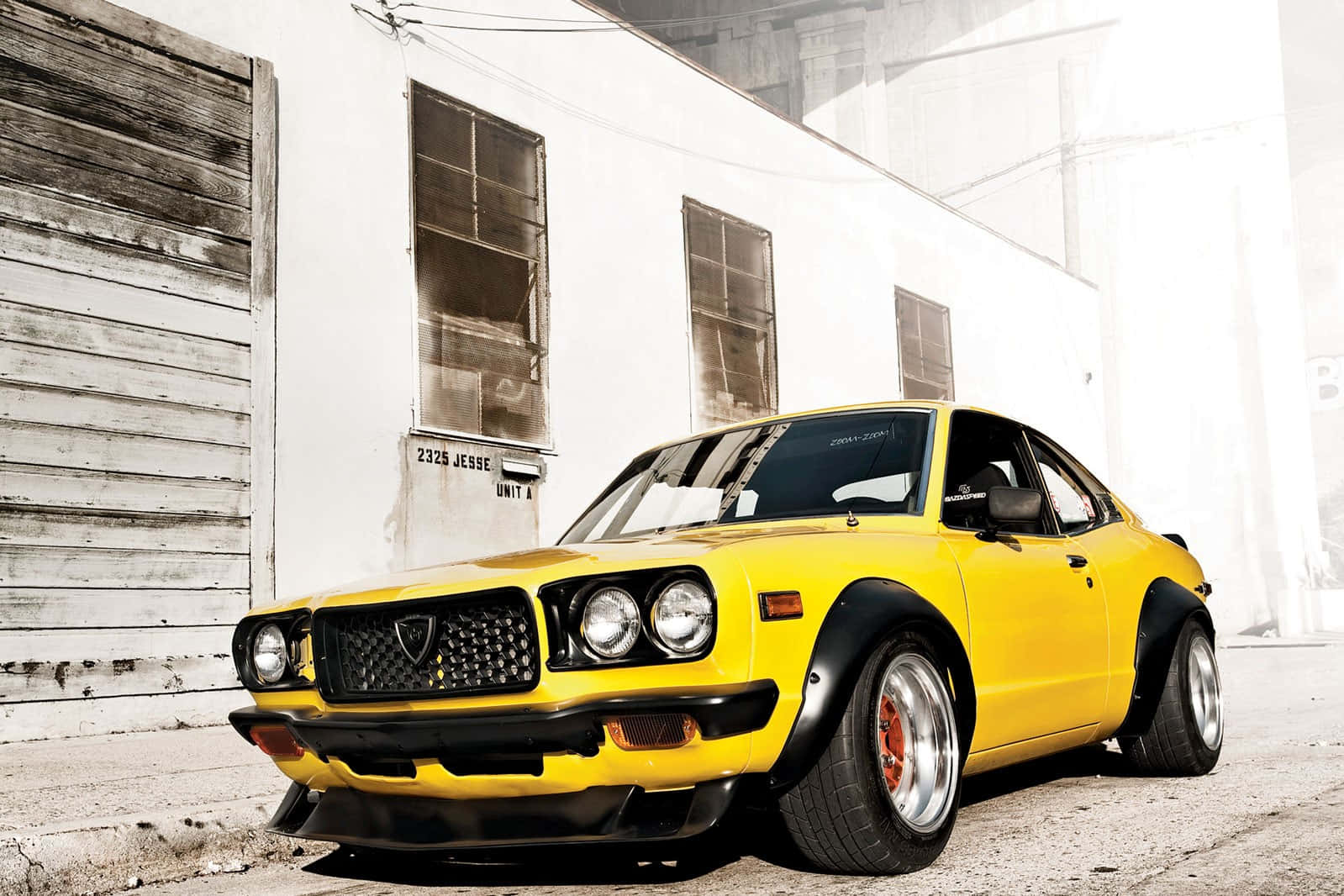 Mazda Rx-3 桌布 壁紙 — 一輛充滿活力的黃色經典跑車，帶有黑色裝飾，停在風化的工業建築前。