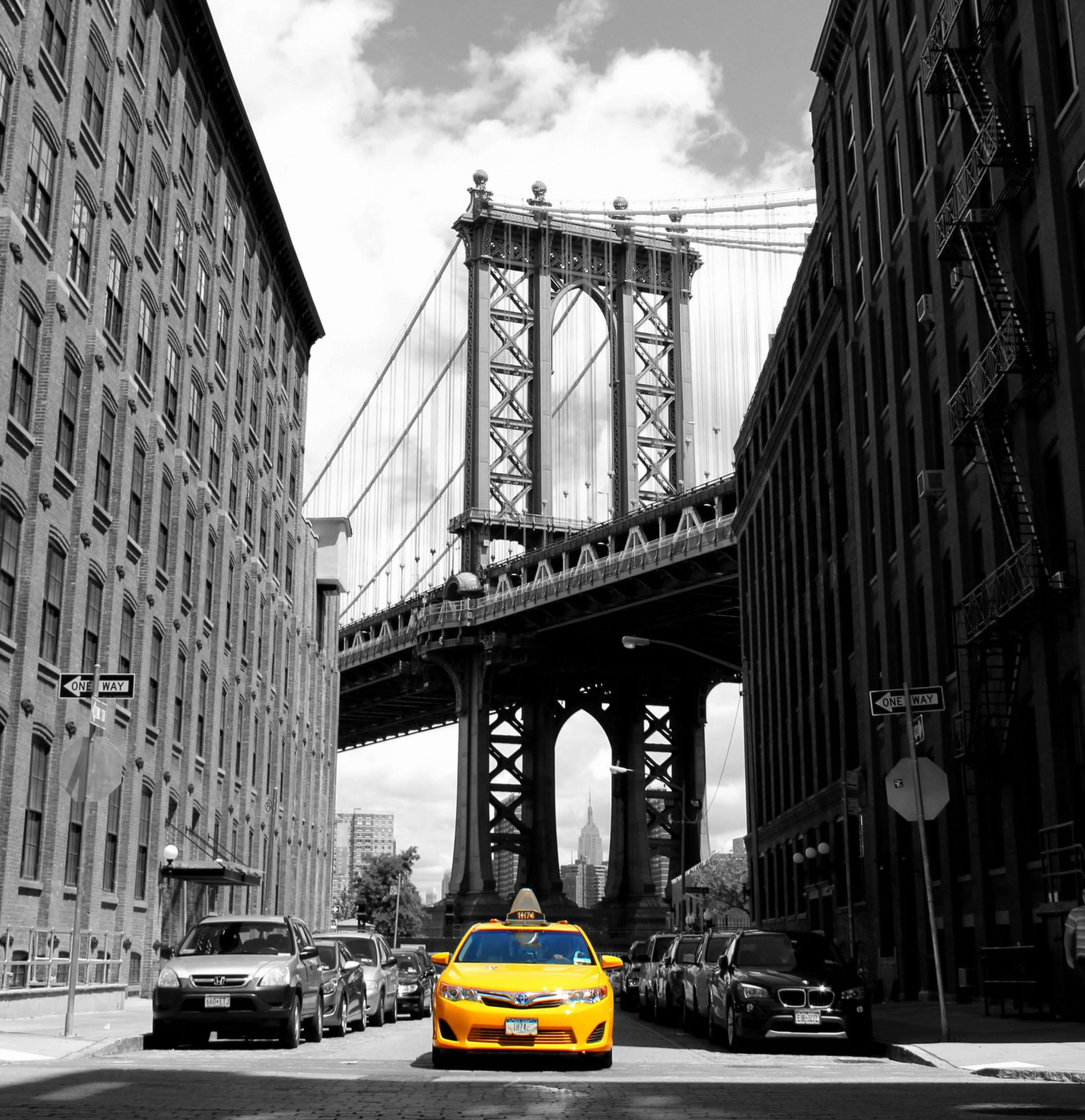 Yellow Cab 桌布 壁紙 — 一輛充滿活力的黃色計程車行駛在狹窄的紐約街道上，兩旁是高聳的磚造建築，曼哈頓大橋以黑白風格聳立在上方。