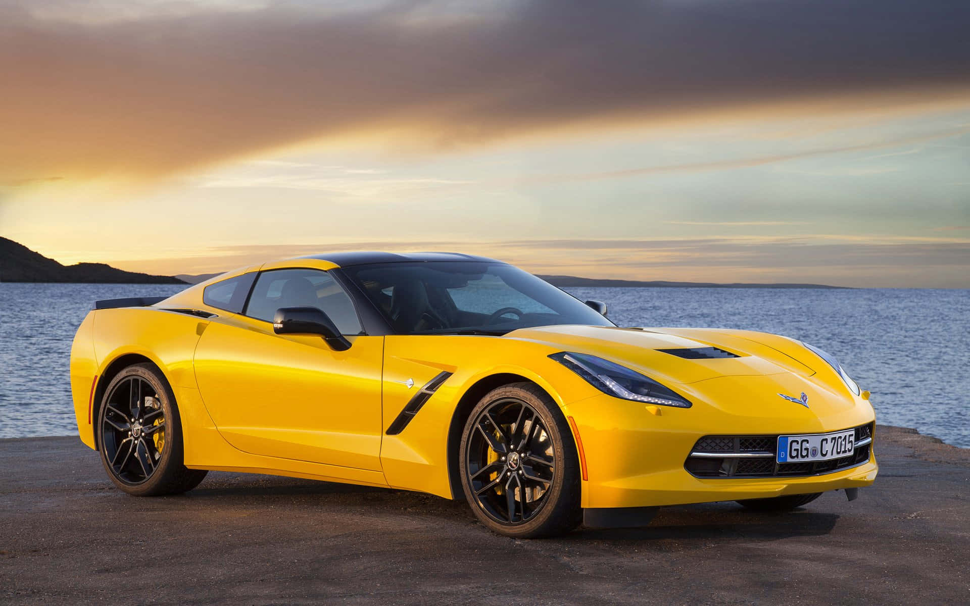 Chevrolet Corvette Stingray 桌布 壁紙 — 一輛充滿活力的黃色跑車在日落時停在水邊，上方有戲劇性的雲層。