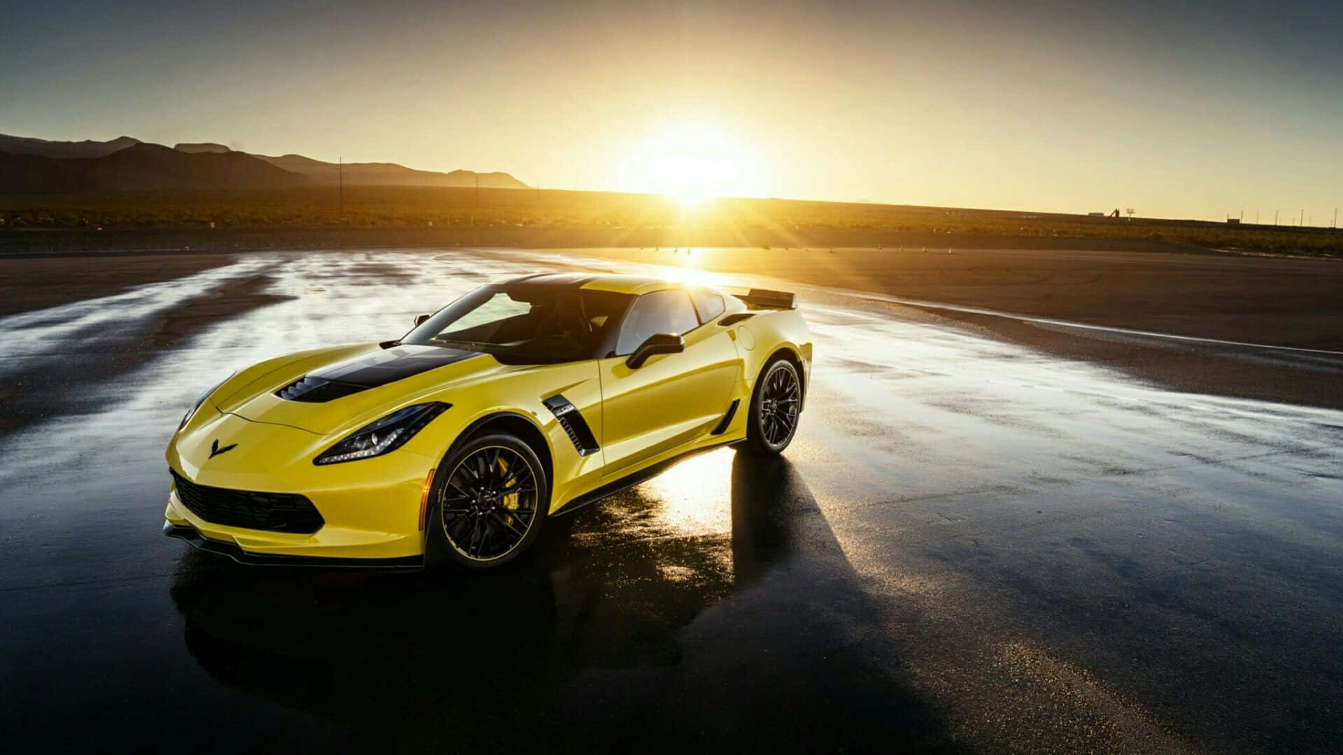 corvette 桌布 壁紙 — 一輛充滿活力的黃色跑車在潮濕的賽道上閃閃發光，夕陽在遠山後落下，投下長長的金色陰影。