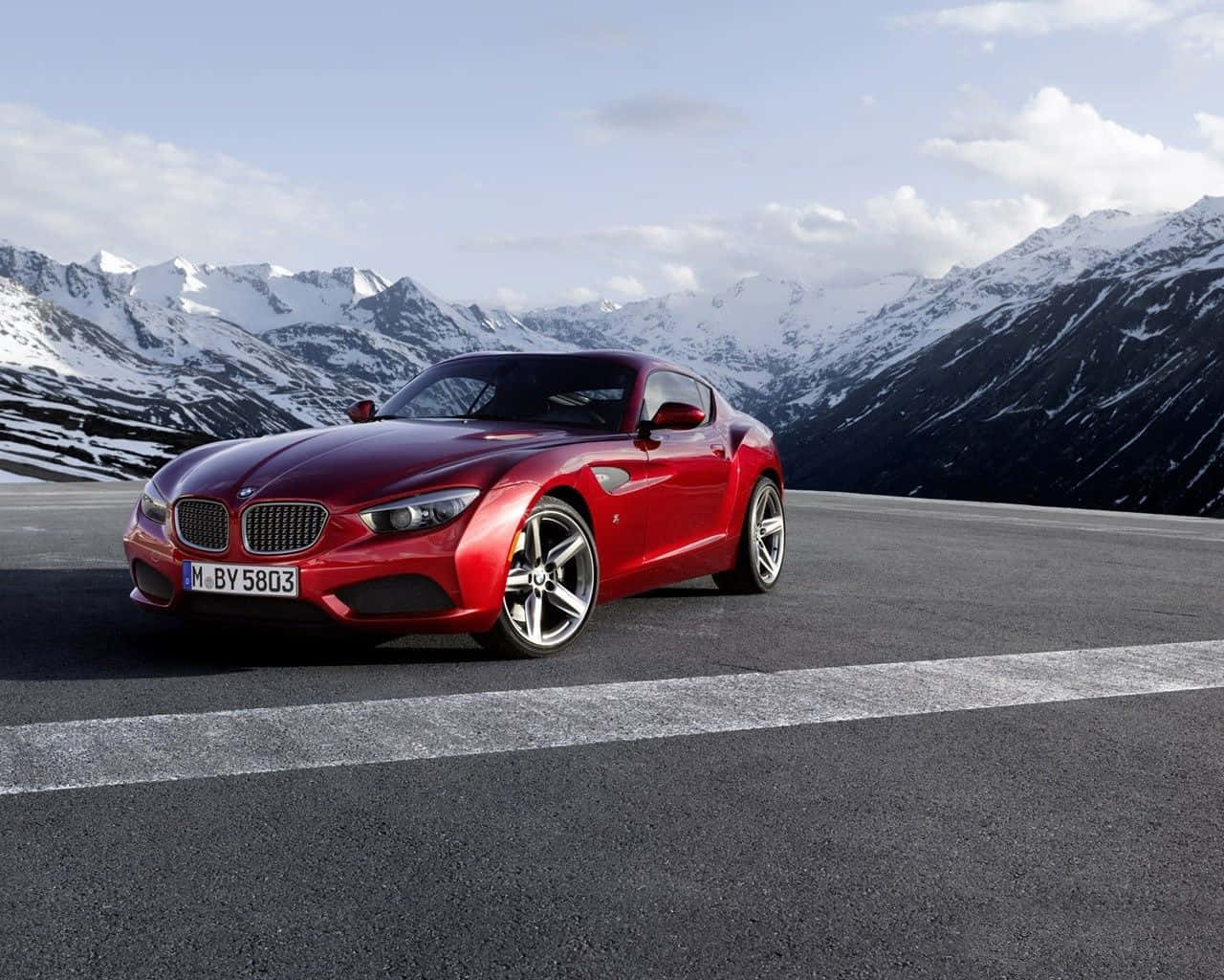 Bmw Z4 桌布 壁紙 — 一輛光亮的紅色BMW Z4跑車停泊在山路上，背景是部分多雲的天空下的積雪山峰。