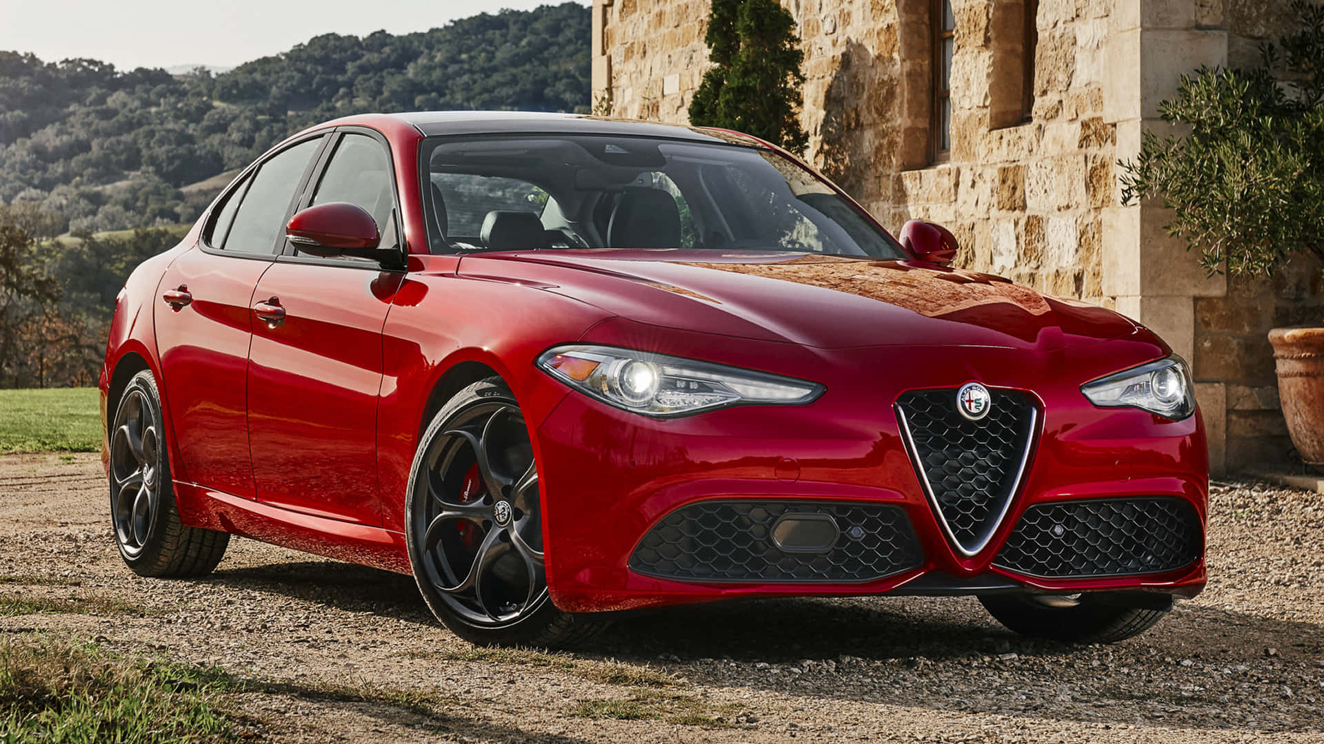 Alfa Romeo Giulia 桌布 壁紙 — 一輛光亮的紅色愛快羅密歐房車停在碎石路上，旁邊是石頭建築，背景有山丘。