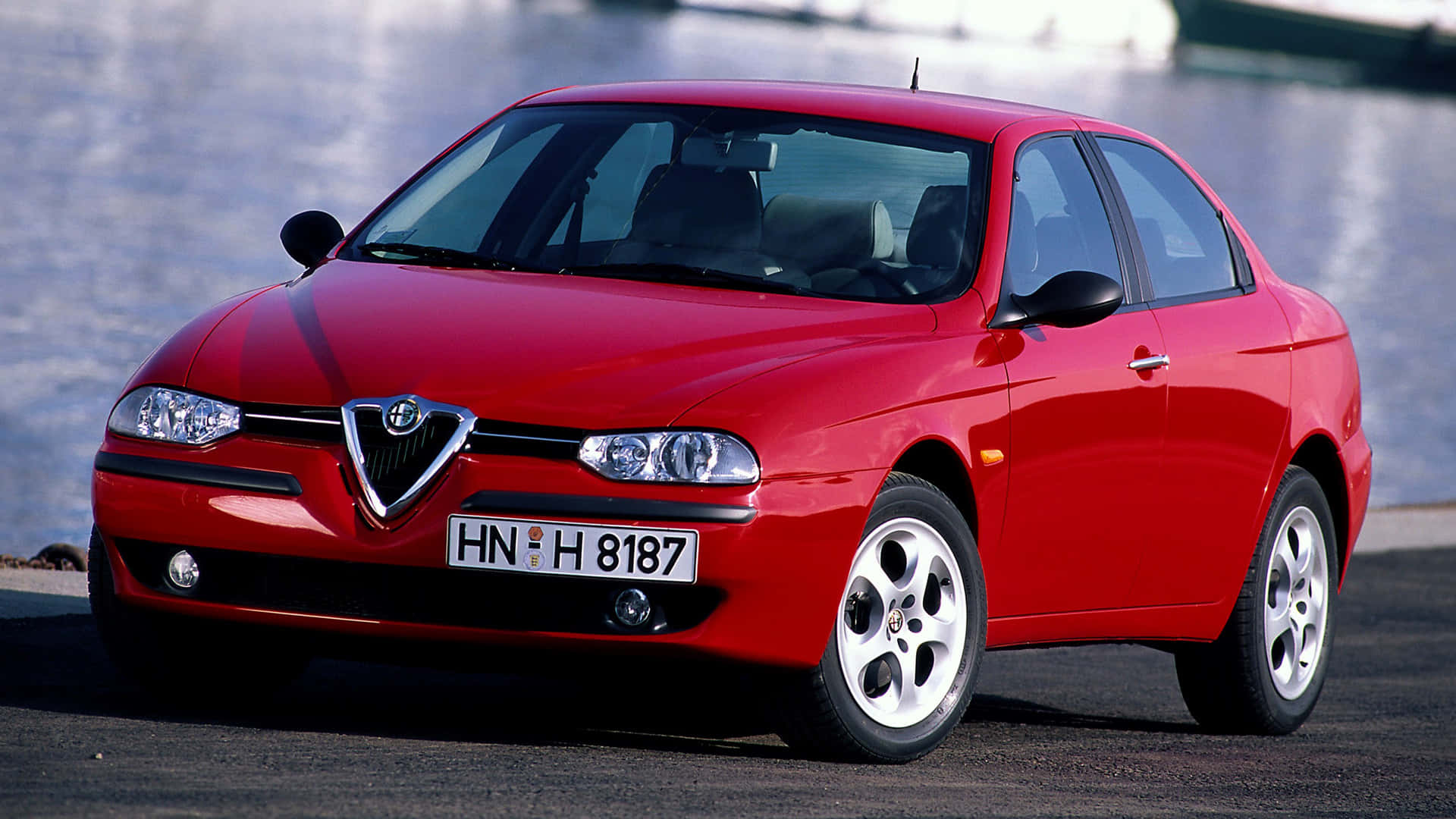 Alfa Romeo 156 桌布 壁紙 — 一輛光亮的紅色愛快羅密歐房車停泊在河道旁，在明亮的日光下，拋光表面閃爍著倒影。