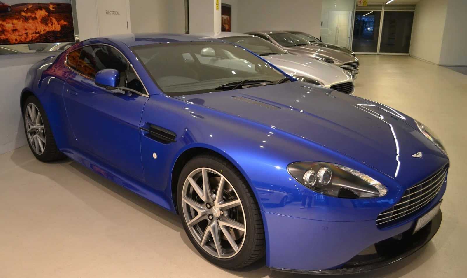 Cobalt Blue 桌布 壁紙 — 一輛光亮的藍色Aston Martin跑車在展廳燈光下閃閃發光，後面可見另一輛銀色汽車。
