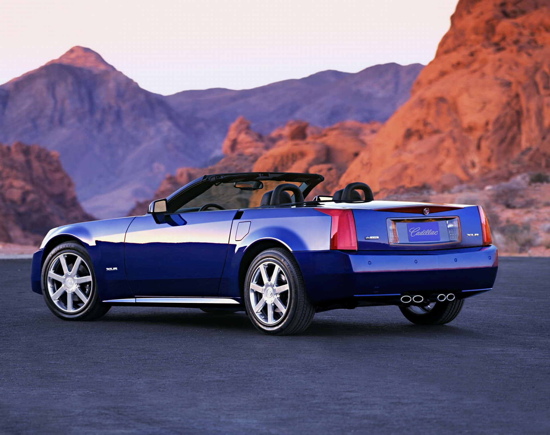 Cadillac Xlr 桌布 壁紙 — 一輛光亮的藍色Cadillac XLR開篷車停在瀝青路上，紅色岩石地貌和紫色山脈在夕陽餘暉中閃耀。