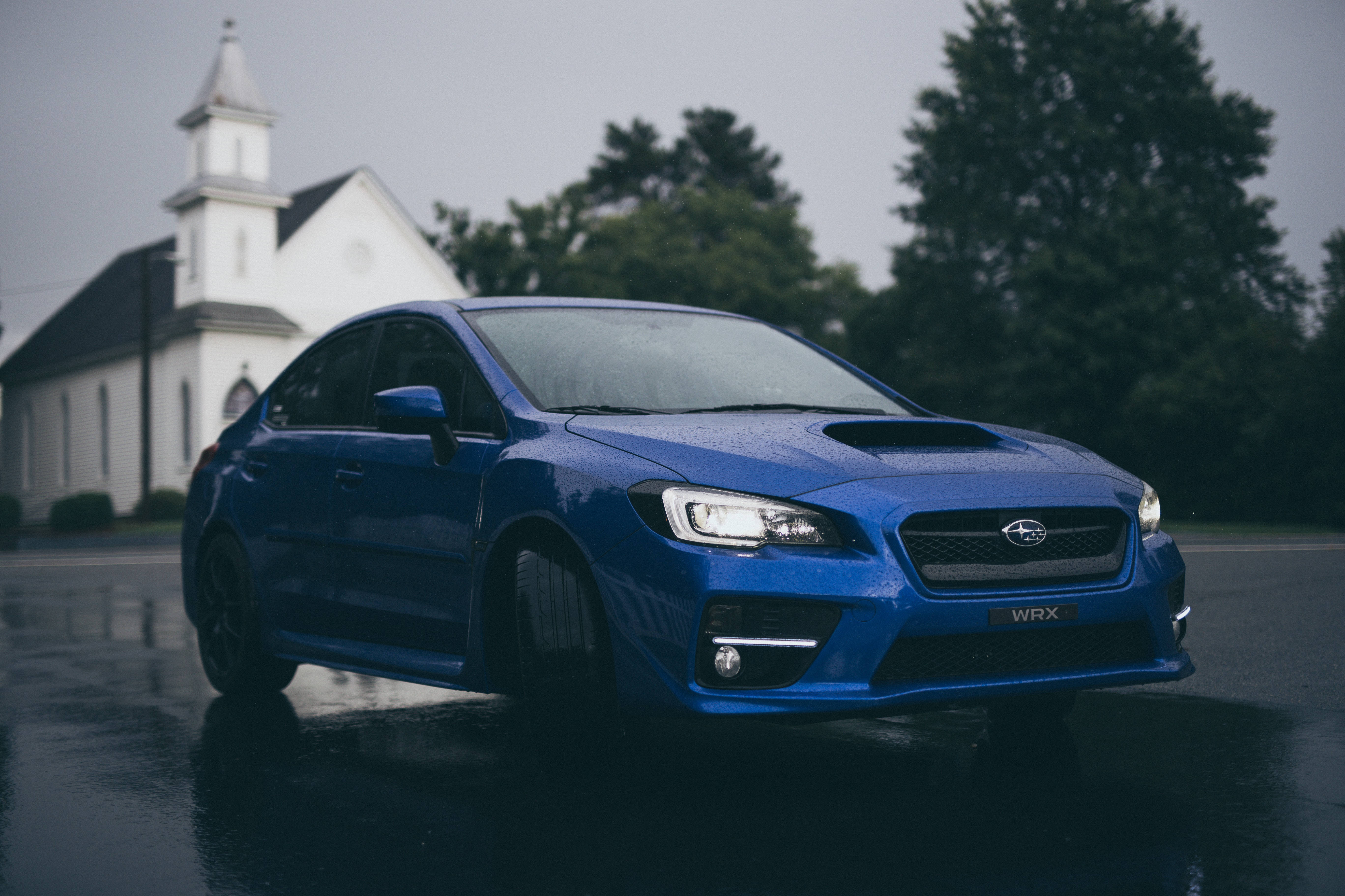 subaru 桌布 壁紙 — 一輛光亮的藍色Subaru WRX房車在潮濕的道路上閃閃發光，背景是霧氣中的白色教堂和樹木。