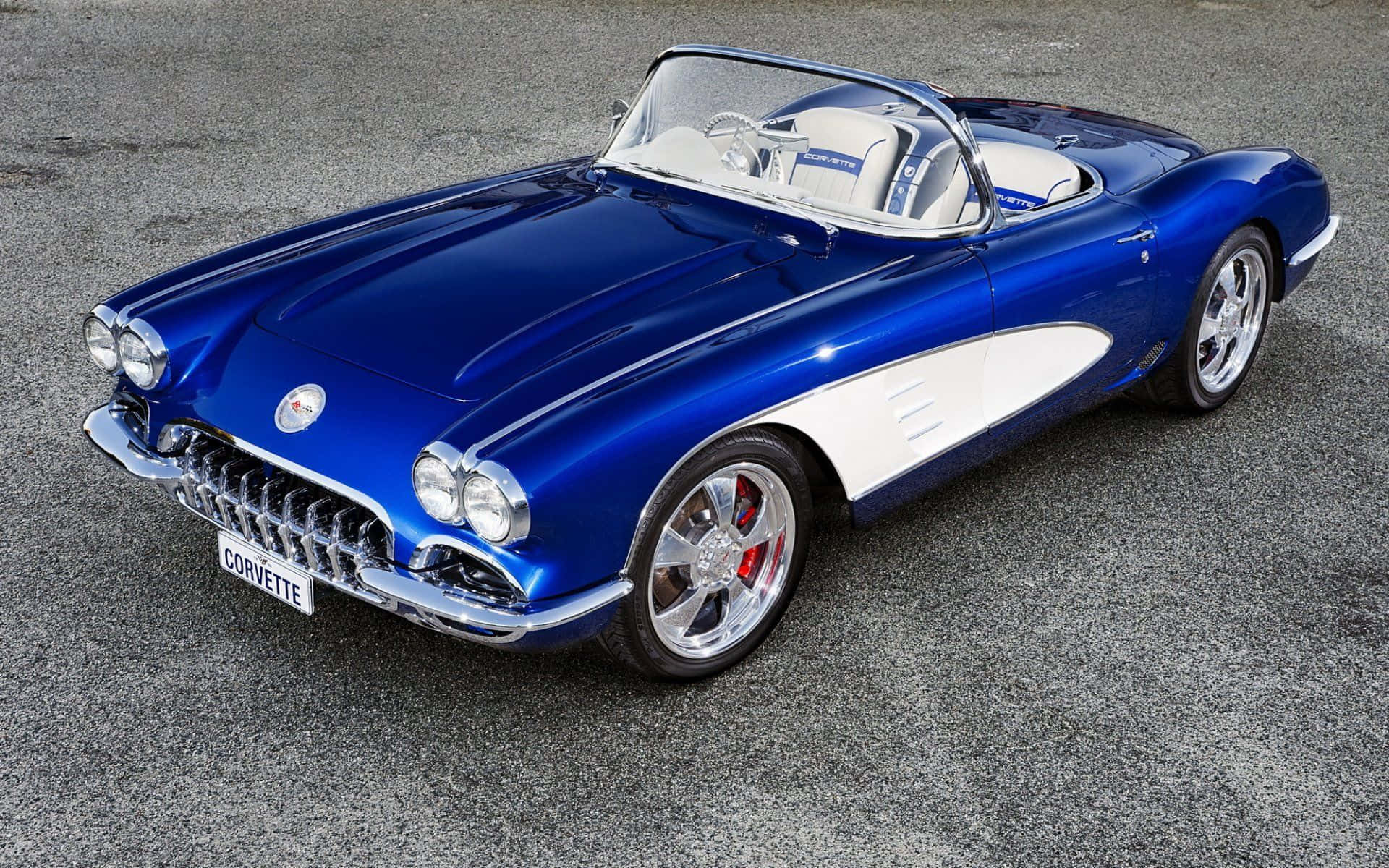 classic 桌布 壁紙 — 一輛光亮的藍色經典Corvette開篷車，帶有白色點綴，停在灰色瀝青上，在明亮的日光下。
