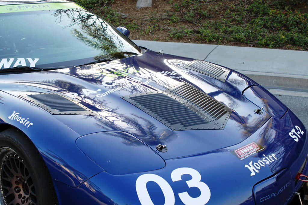 Car Hood 桌布 壁紙 — 一輛光亮的藍色賽車，編號03，帶有Hoosier貼紙，在引擎蓋上反射出附近的樹木，旁邊是人行道。