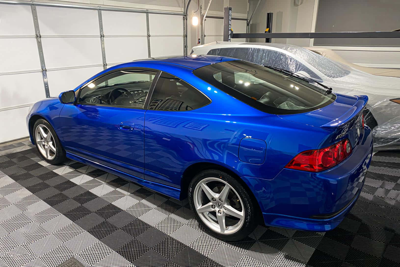 Acura Rsx 桌布 壁紙 — 一輛光亮的藍色運動型轎跑車停在格子車庫地板上，後面有一輛用塑料覆蓋的白色汽車。