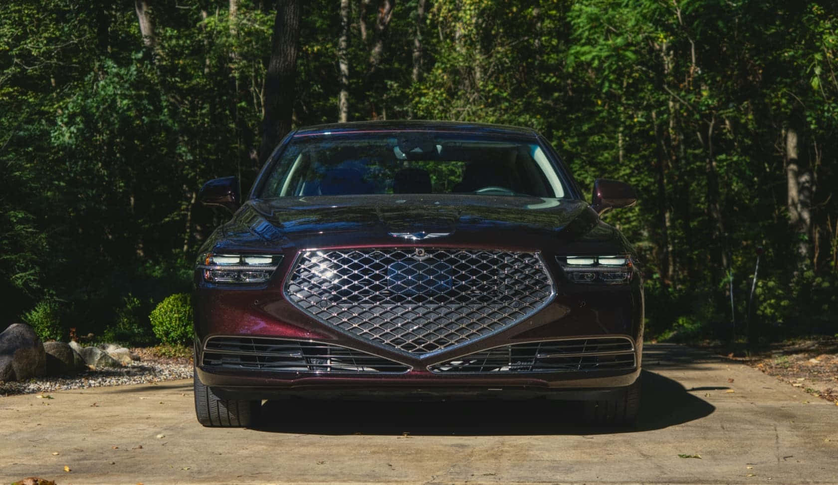 Genesis G90 桌布 壁紙 — 一輛光澤的栗色豪華轎車，帶有大型鍍鉻格柵，停在混凝土車道上，周圍是茂密的綠色森林。