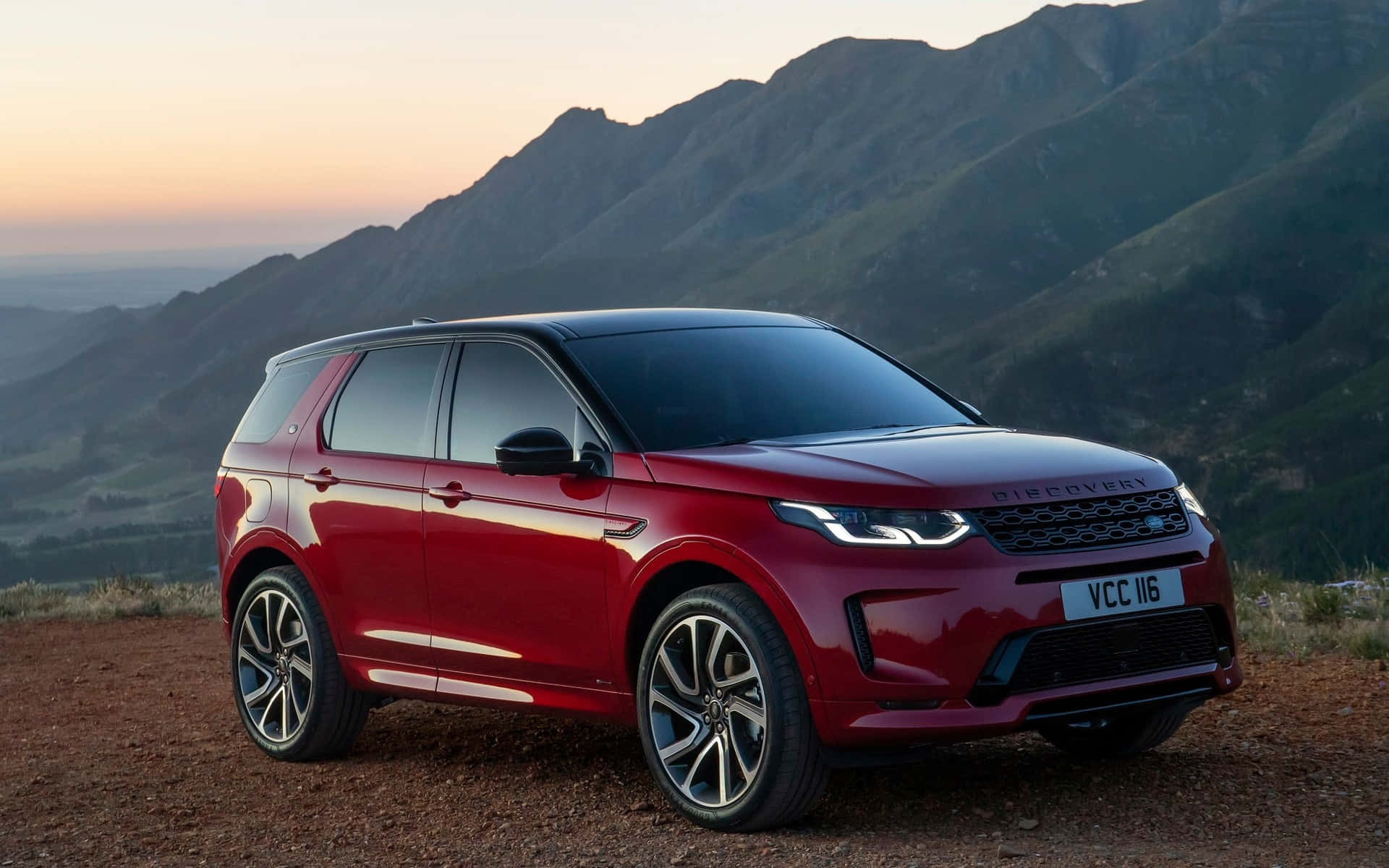 Land Rover Discovery 桌布 壁紙 — 一輛光澤的紅色 Land Rover Discovery 停泊在泥土觀景點，俯瞰著沐浴在溫暖日落光線中的連綿山脈。