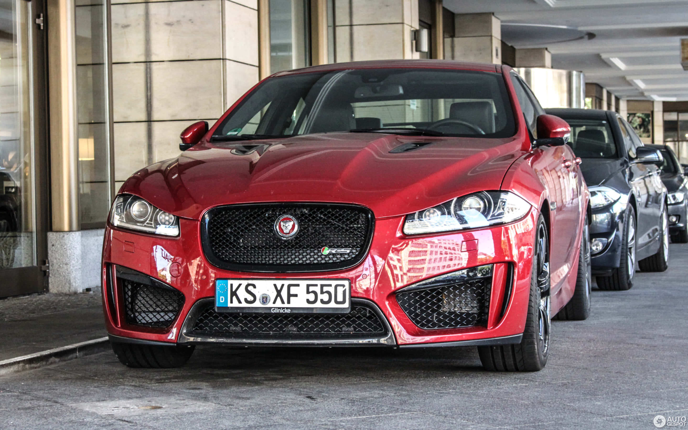 Jaguar Xfr 桌布 壁紙 — 一輛光澤的紅色Jaguar XF 550運動轎車停放在一座具有反射表面的現代建築下，附近還有其他車輛。