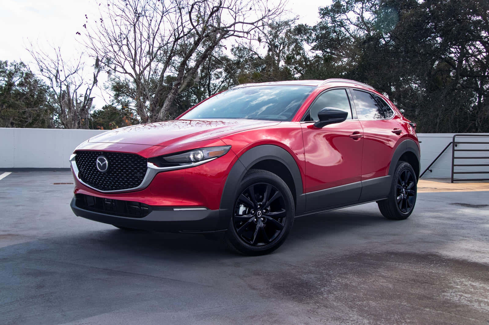 Mazda Cx-30 桌布 壁紙 — 一輛光澤的紅色Mazda CX-30 SUV，帶有黑色飾邊，停在混凝土停車場，周圍是落葉樹。