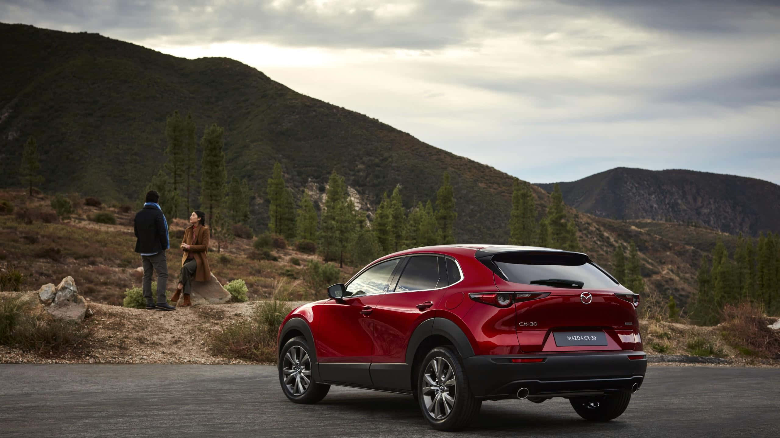Mazda Cx-30 桌布 壁紙 — 一輛光澤的紅色Mazda CX-30 SUV停泊在鋪砌道路上，附近站著一對情侶，背景是起伏的山丘和稀疏的樹木，天空多雲。