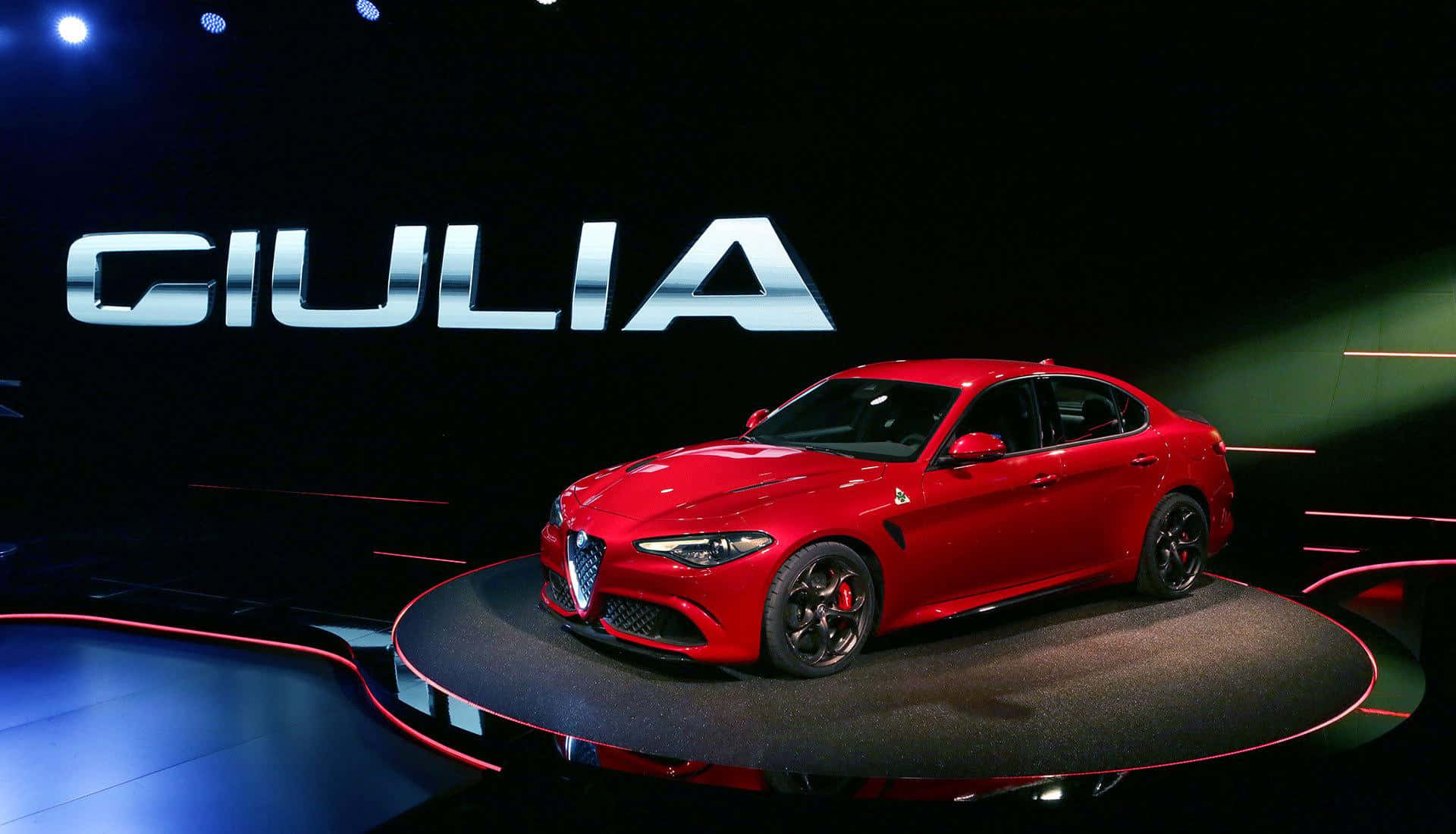 Alfa Romeo Giulia 桌布 壁紙 — 一輛光澤的紅色愛快羅密歐Giulia轎車在圓形舞台上於聚光燈下閃耀，背後有大型銀色字母「GIULIA」。