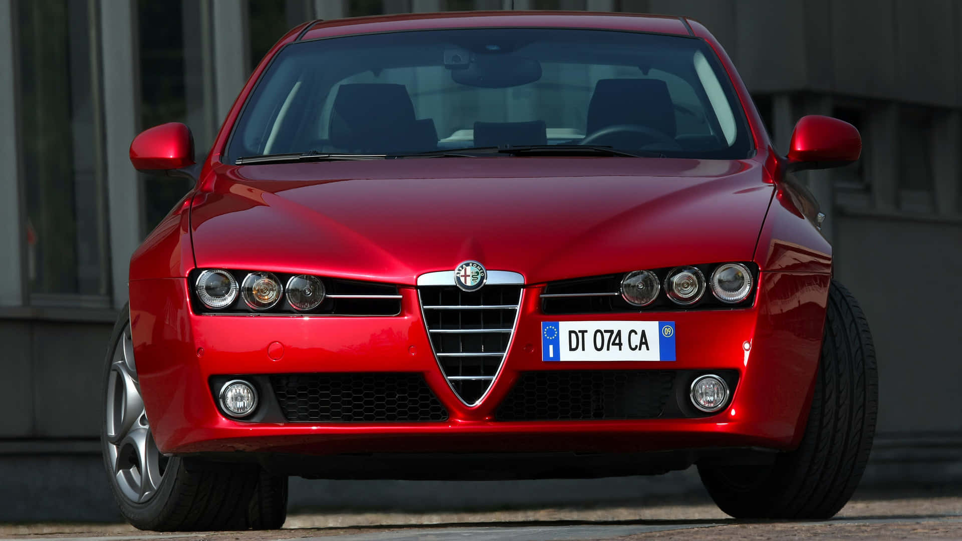 Alfa Romeo 159 桌布 壁紙 — 一輛光澤的紅色愛快羅密歐跑車，帶有獨特的格柵和圓形頭燈，停泊在城市的街道上。
