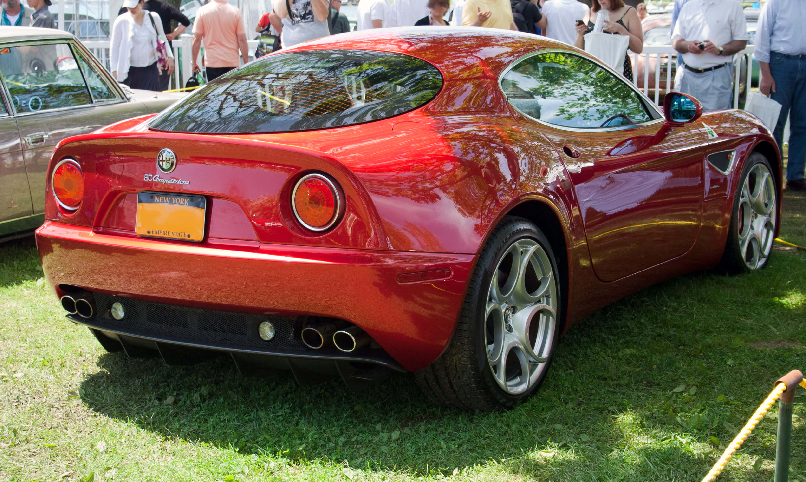 Alfa Romeo 8c Competizione 桌布 壁紙 — 一輛光澤的紅色愛快羅密歐跑車在明亮的陽光下，在附近的草地上閃閃發光，旁邊有觀眾。