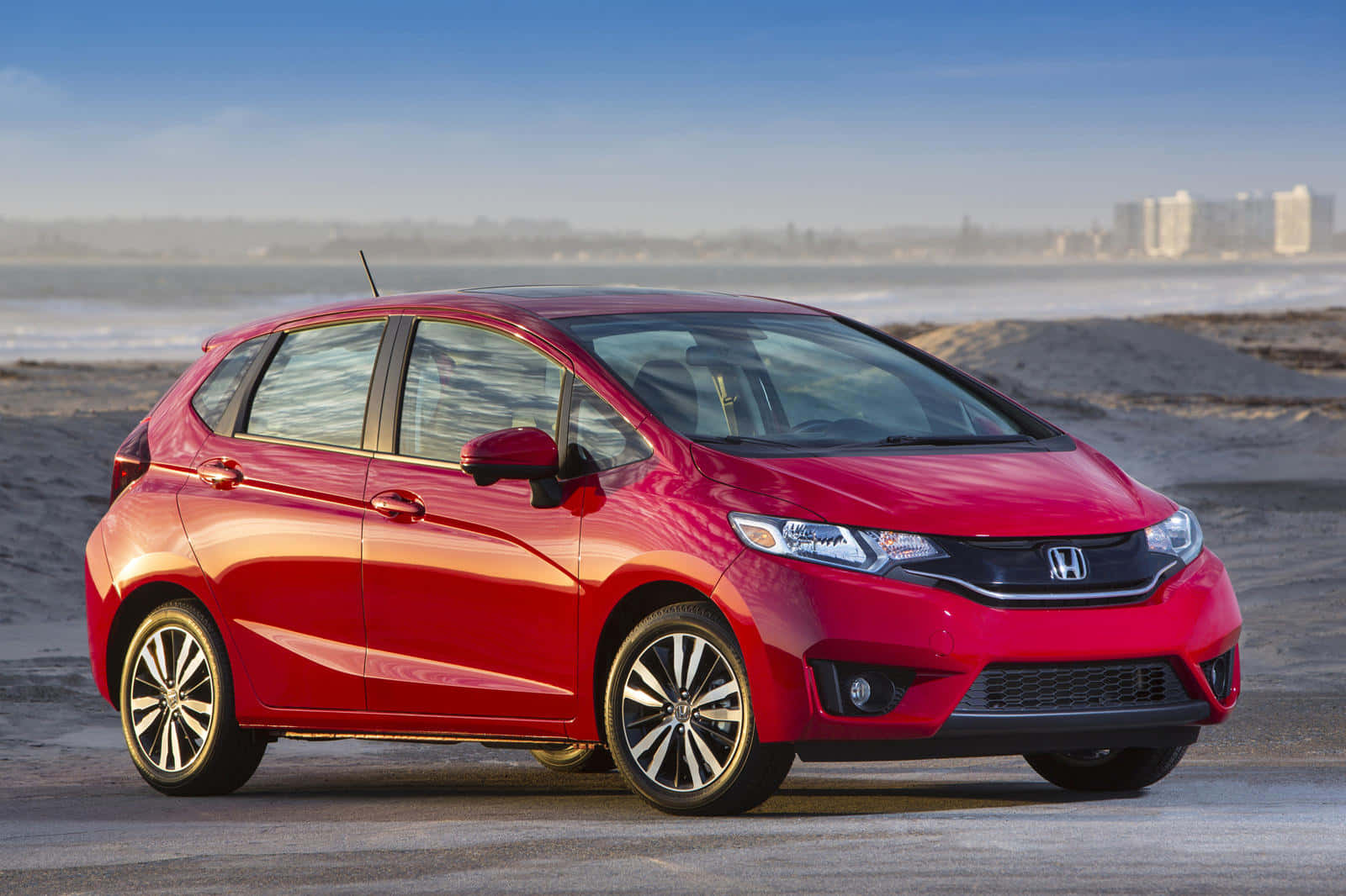 Honda Fit 桌布 壁紙 — 一輛光澤的紅色本田Fit停泊在沙灘上，海浪和遠處的城市天際線在晴朗的藍天下。