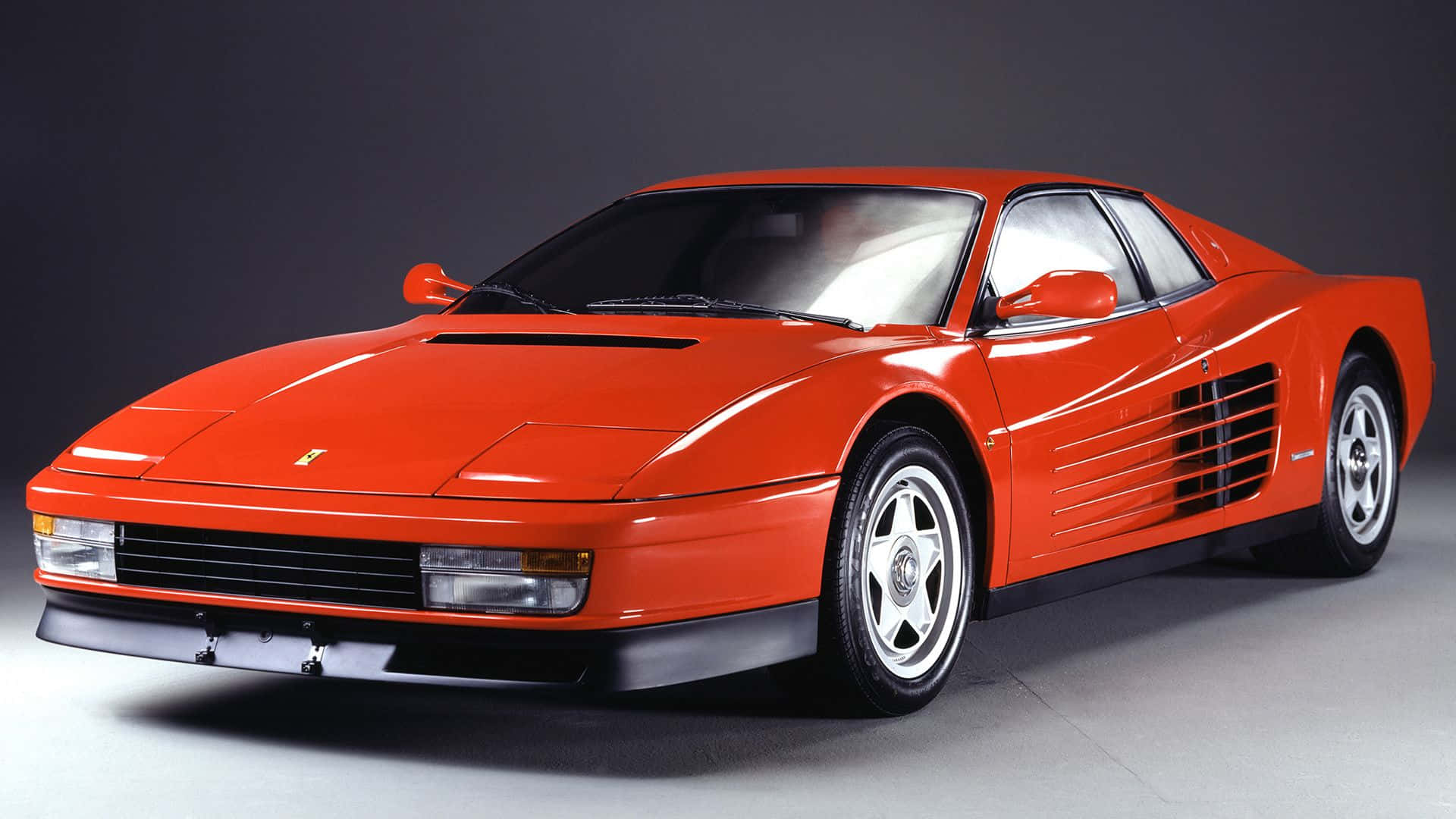 Ferrari Testarossa 桌布 壁紙 — 一輛光澤的紅色法拉利 Testarossa 跑車，帶有標誌性的側面進氣口和銀色輪圈，在深色工作室背景下擺拍。