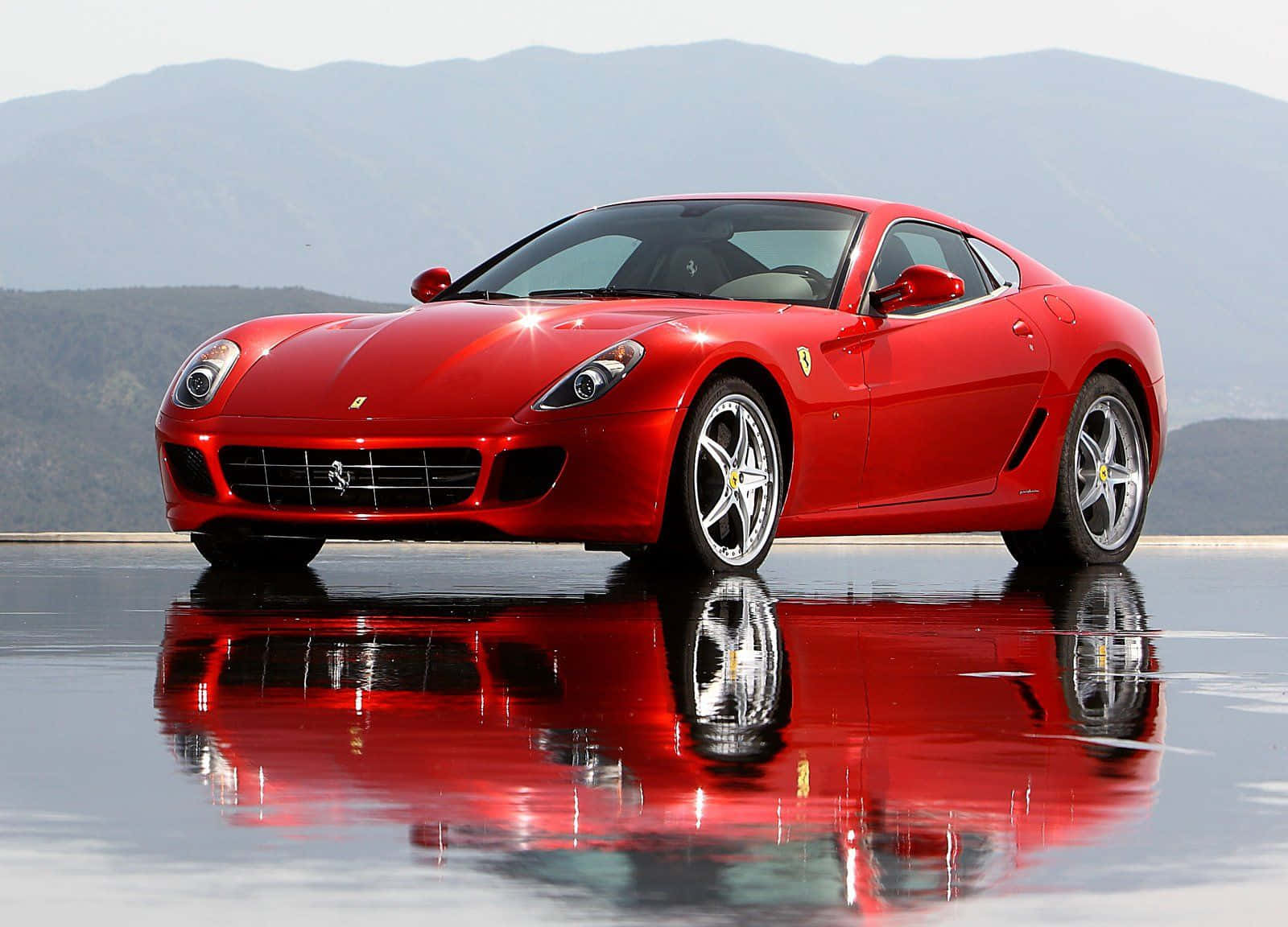 Ferrari 599 Gtb Fiorano 桌布 壁紙 — 一輛光澤的紅色法拉利跑車在濕滑路面上閃耀，背景是山脈，倒影完美。