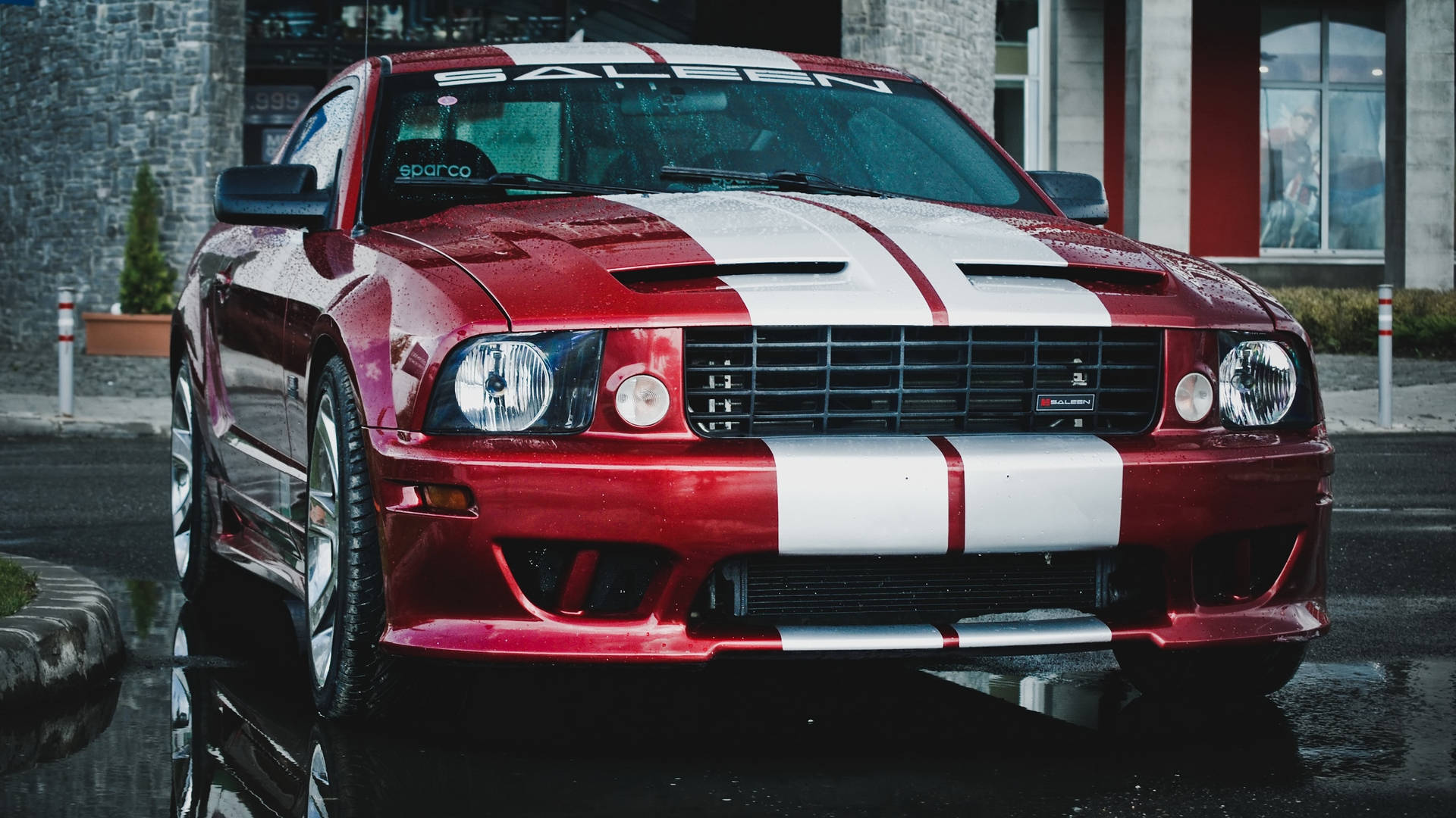 race car 桌布 壁紙 — 一輛光澤的紅色福特野馬，帶有白色賽車條紋，停在潮濕的城市街道上，反射出其大膽的設計。