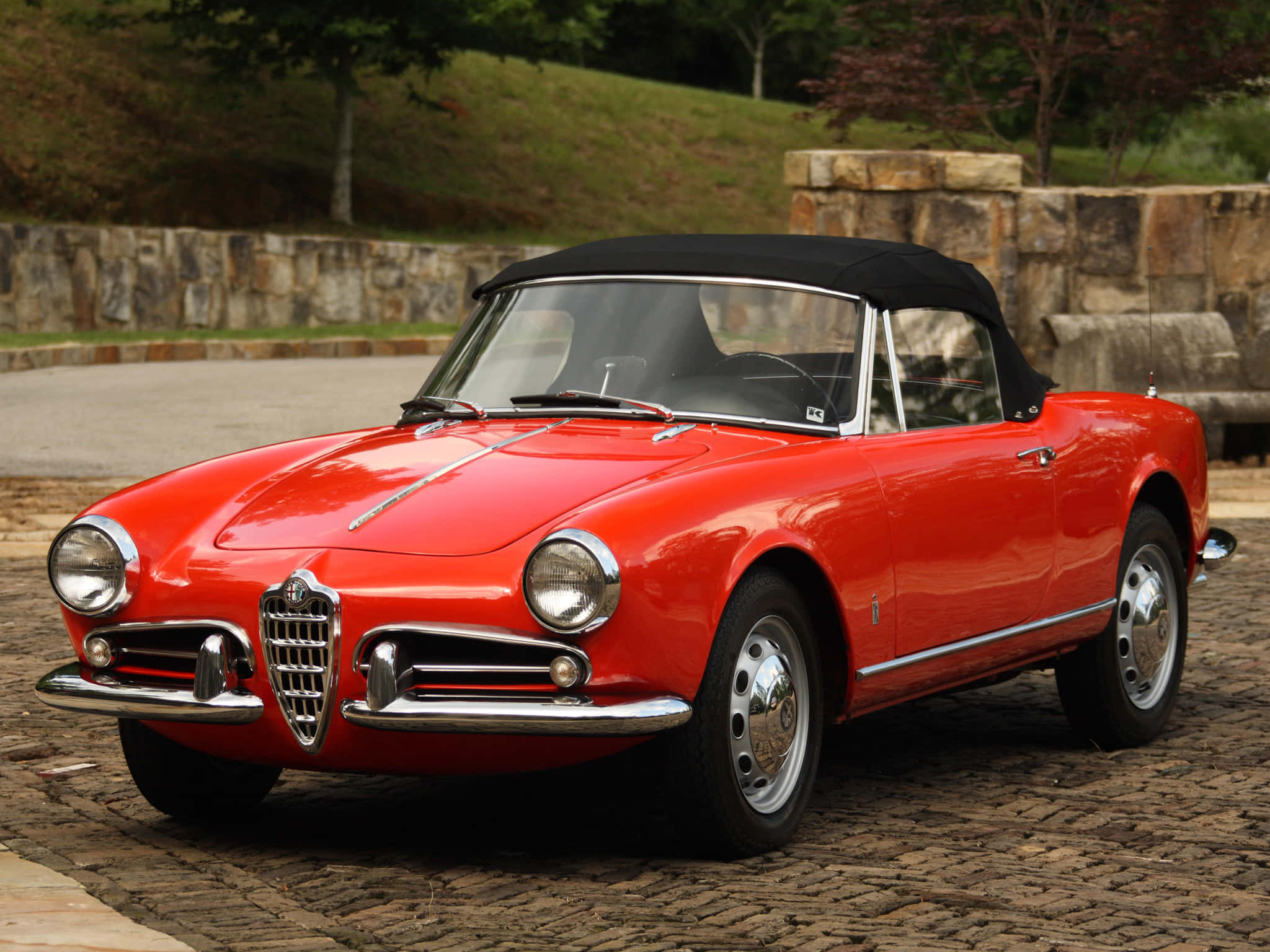 Alfa Romeo Spider 桌布 壁紙 — 一輛光澤的紅色經典愛快羅密歐Spider開篷車，配黑色軟頂，停泊在石牆和綠植旁的鵝卵石車道上。
