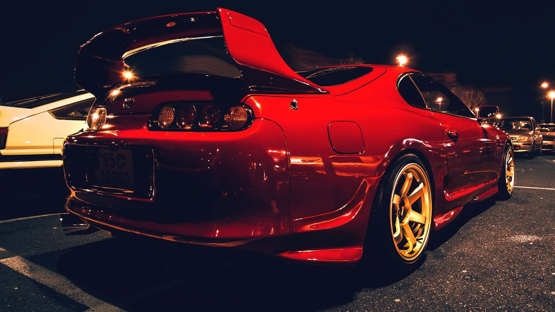 Supra 桌布 壁紙 — 一輛光澤的紅色跑車，配備金色輪圈和升高尾翼，夜晚停在有照明的停車場。