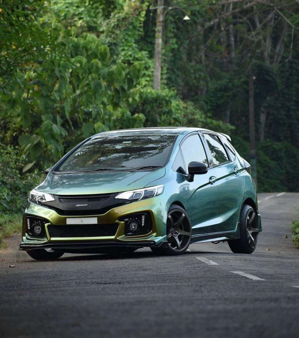 Honda Fit 桌布 壁紙 — 一輛光澤的綠色本田Jazz，配黑色輪圈，停在森林環繞的道路上，在柔和日光下。