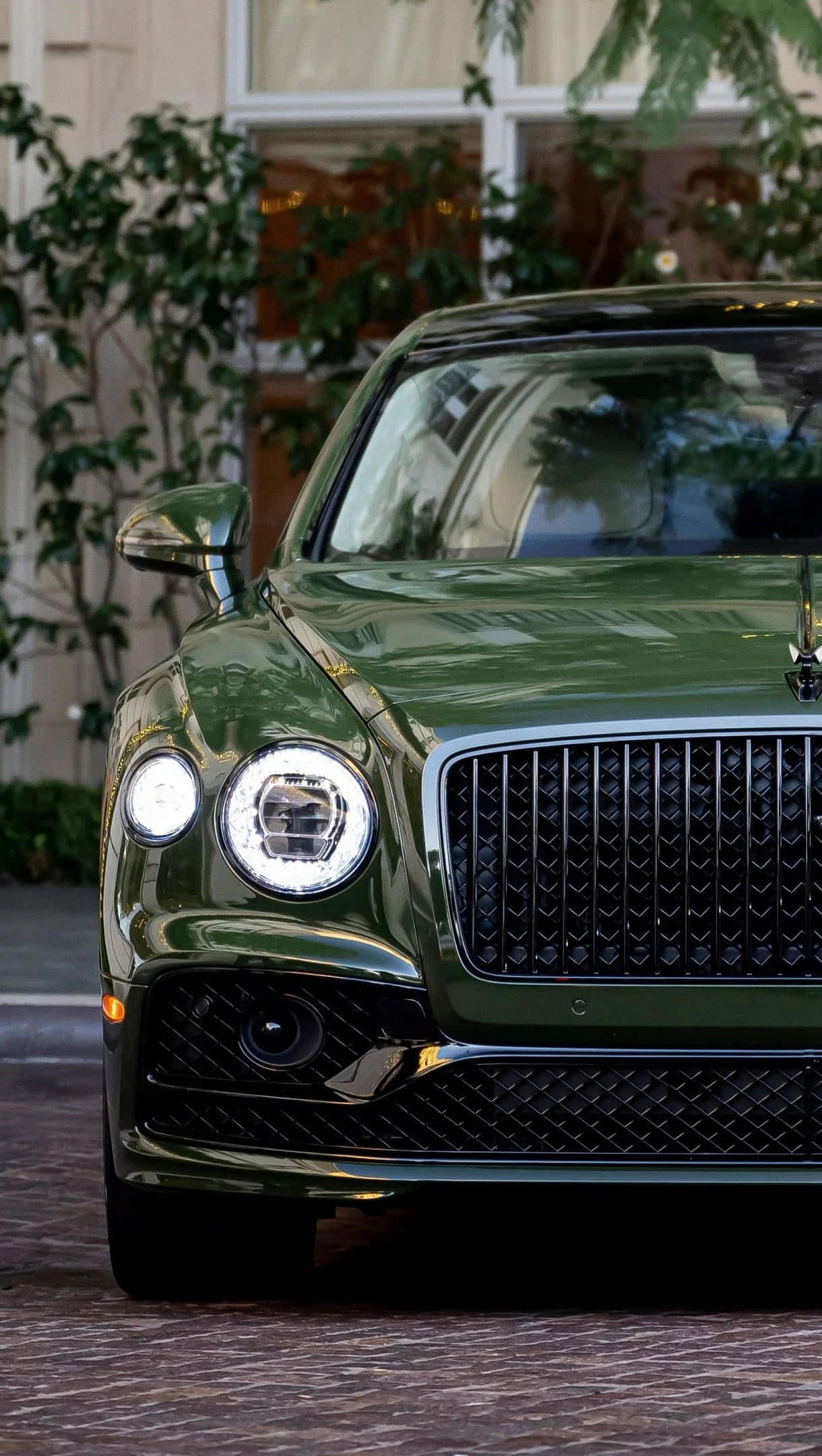 Bentley Flying Spur 桌布 壁紙 — 一輛光澤的綠色賓利 Bentayga 在日光下閃耀，其標誌性格柵和圓形頭燈反射出茂密的花園背景。