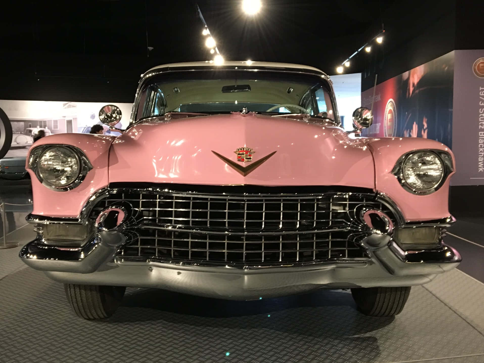 Pink Cadillac 桌布 壁紙 — 一輛光澤粉紅色的1950年代凱迪拉克，帶有鍍鉻格柵和圓形頭燈，在明亮的展覽燈光下閃閃發光。