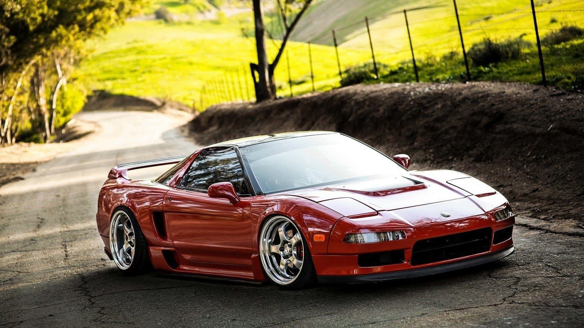 Acura Nsx 桌布 壁紙 — 一輛光澤紅色跑車配備訂製輪圈，停泊在蜿蜒的鄉村道路上，周圍環繞著綠色山丘和樹木。