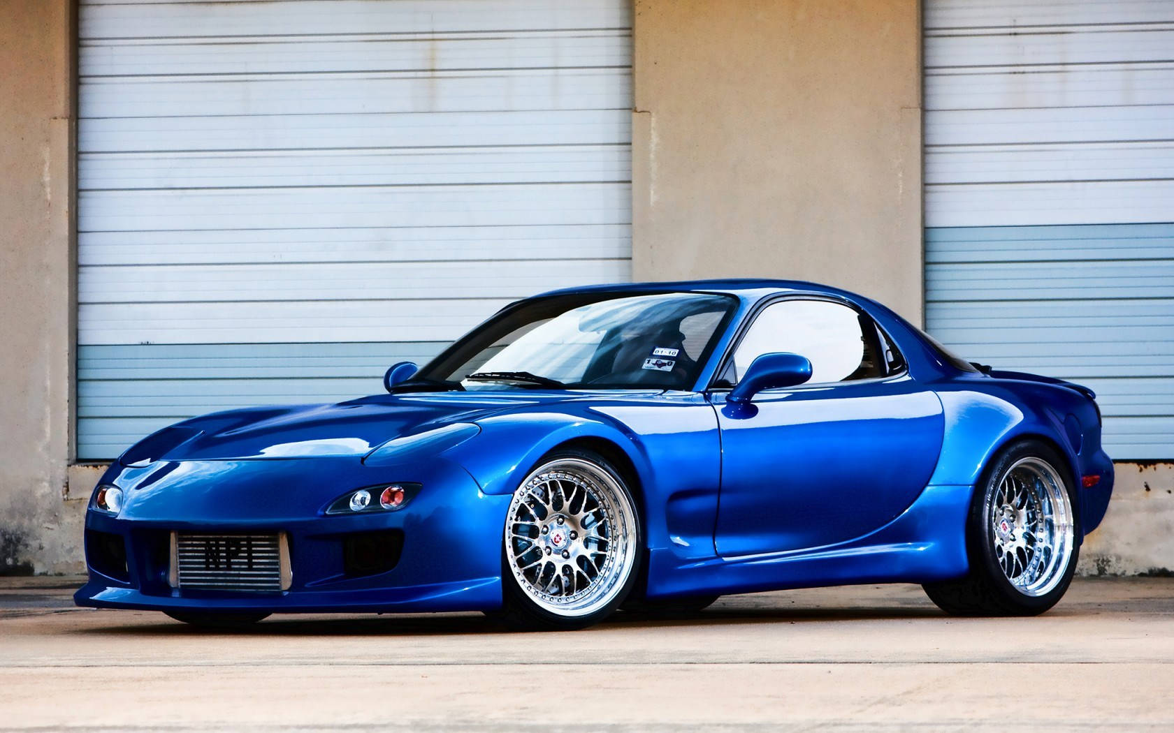 Mazda 桌布 壁紙 — 一輛光澤藍色萬事得RX-7跑車配備定制輪圈，停泊在波紋金屬車庫門前。