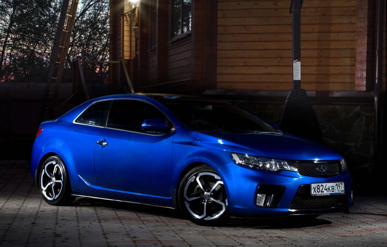 Kia Forte 桌布 壁紙 — 一輛光澤藍色運動型轎跑車在黃昏時停泊在一間帶有溫暖環境照明的木屋旁。