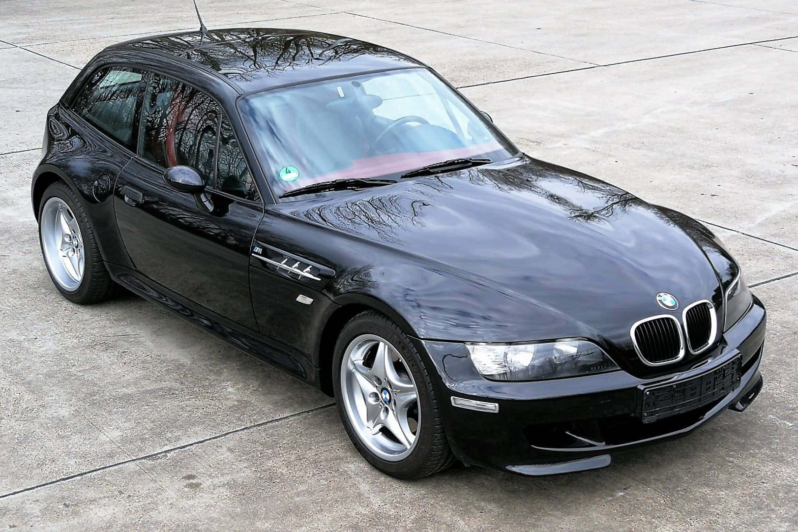 Bmw Z3 桌布 壁紙 — 一輛光澤黑色BMW Z3跑車停在混凝土表面上，拋光引擎蓋上閃爍著樹木的倒影。
