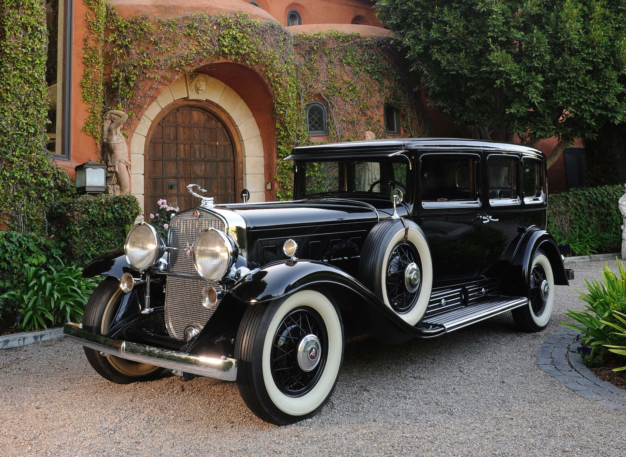 Cadillac Fleetwood 桌布 壁紙 — 一輛光澤黑色復古轎車配白色側壁輪胎，優雅地停在碎石車道上，前方是一座爬滿藤蔓的赤陶豪宅。